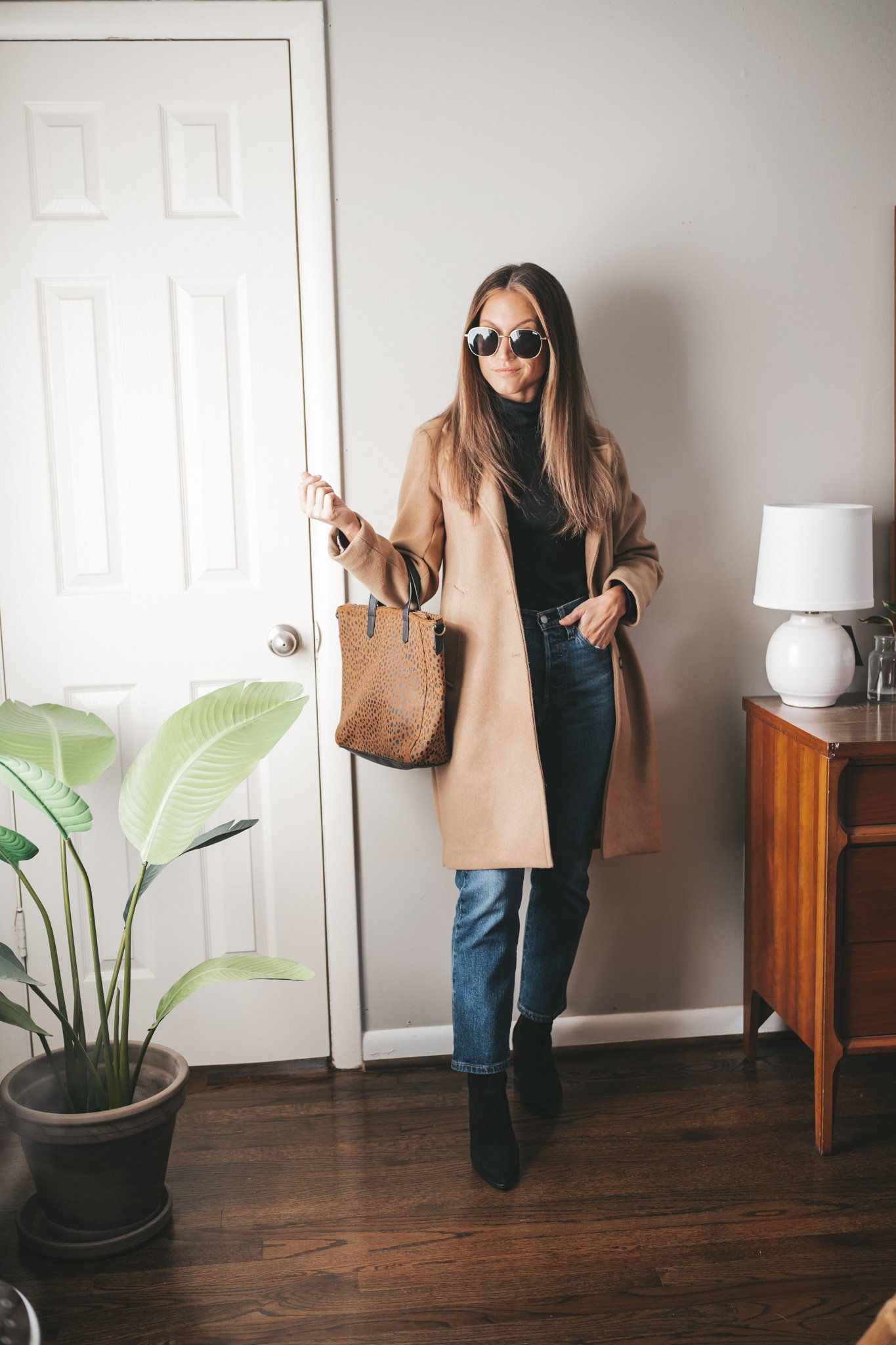 winter capsule wardrobe outfit with a black turtleneck, mid-wash denim, black ankle boots, a camel coat, leopard print tote bag and gold frame oversized sunglasses