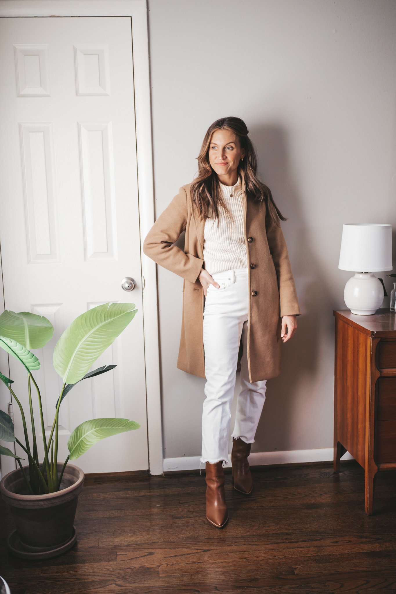 a camel coat outfit idea with a cream colored sweater, white straight leg denim, and brown heeled ankle boots 