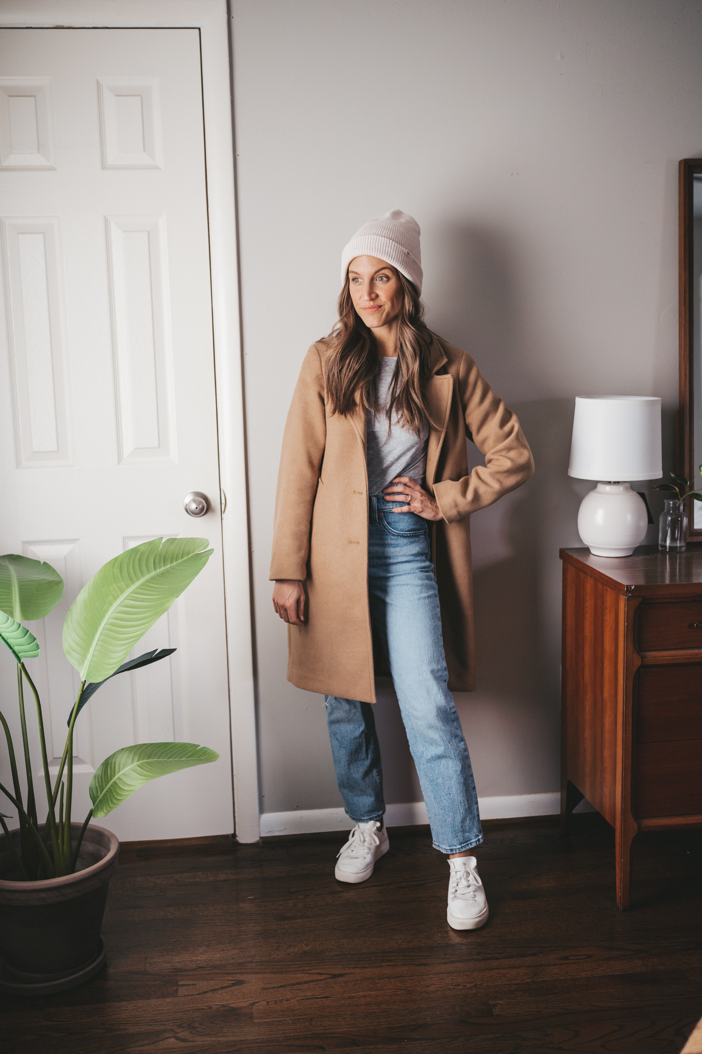 camel coat outfit idea with a gray-long sleeved tee, light wash denim, white sneakers, and a cream colored beanie