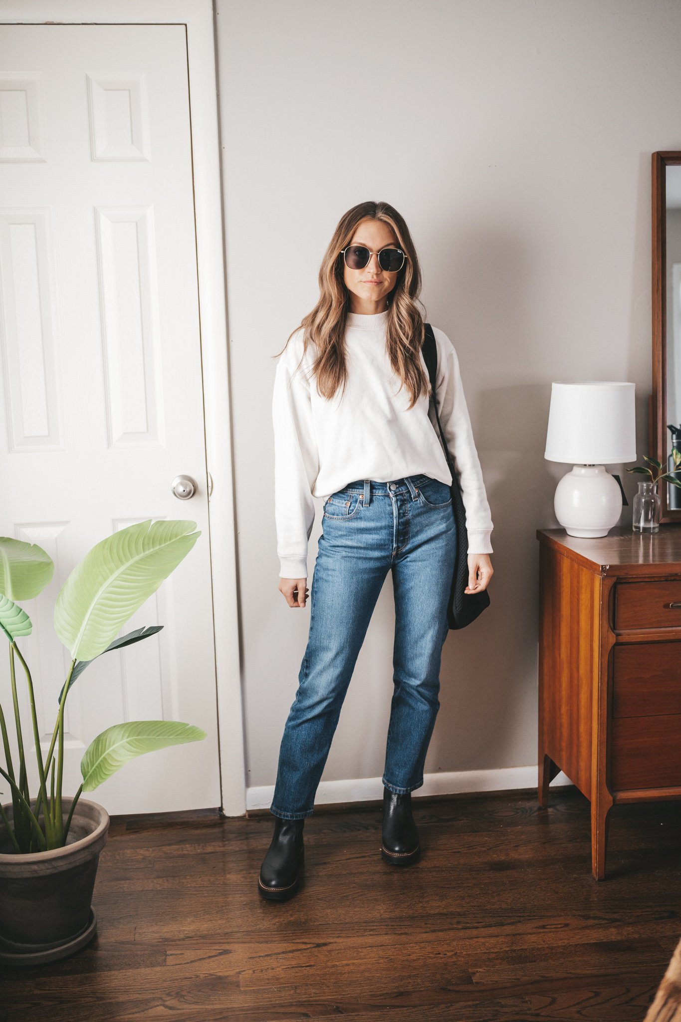 winter capsule wardrobe outfit idea with straight leg dark wash denim, cream colored sweatshirt, black lug-sole chelsea boots, black tote bag and gold frame oversized sunglasses
