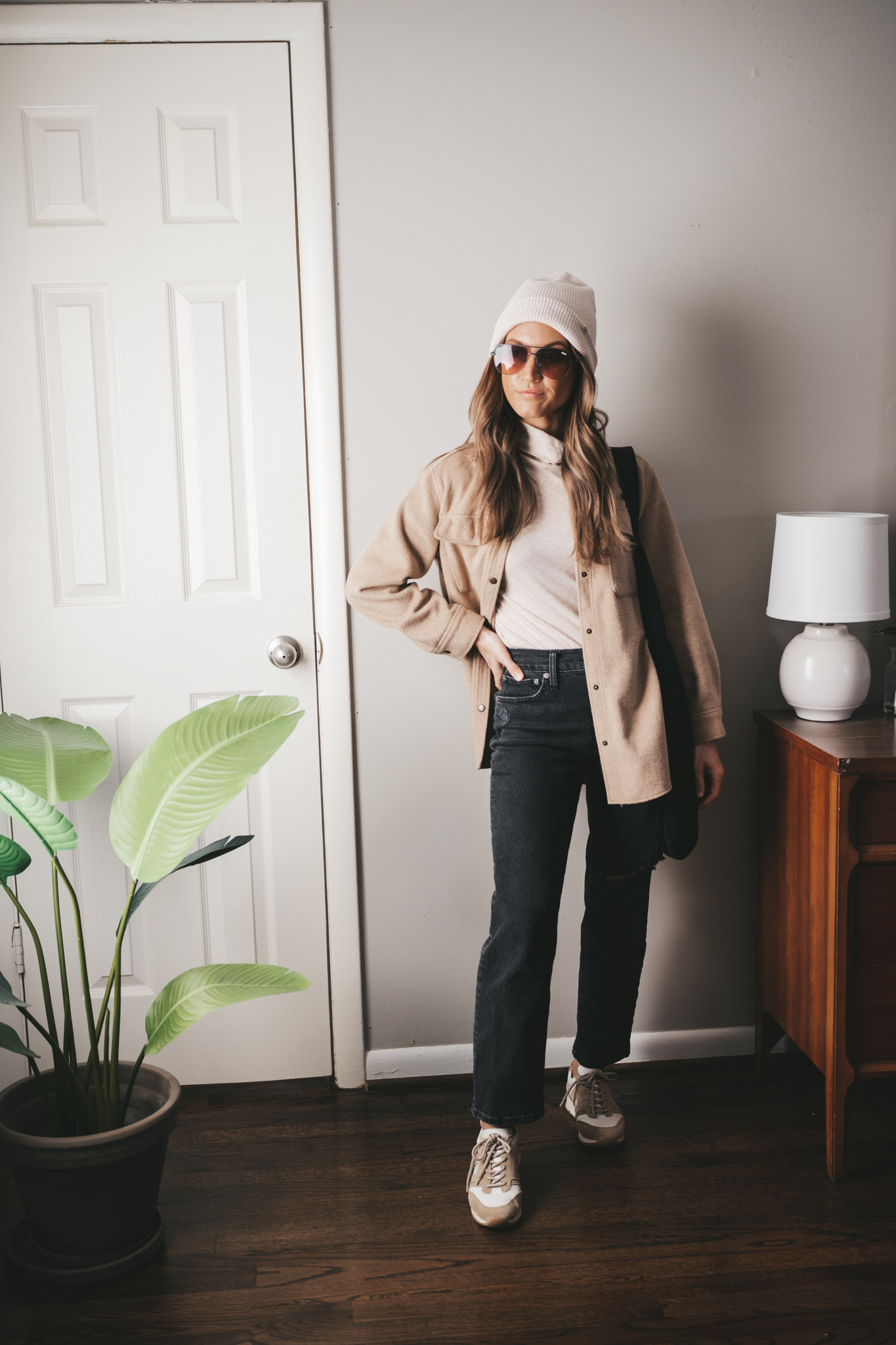 winter capsule wardrobe outfit with a turtleneck, tan shacket, black denim, white and tan sneakers, a cream colored beanie and oversized aviator sunglasses