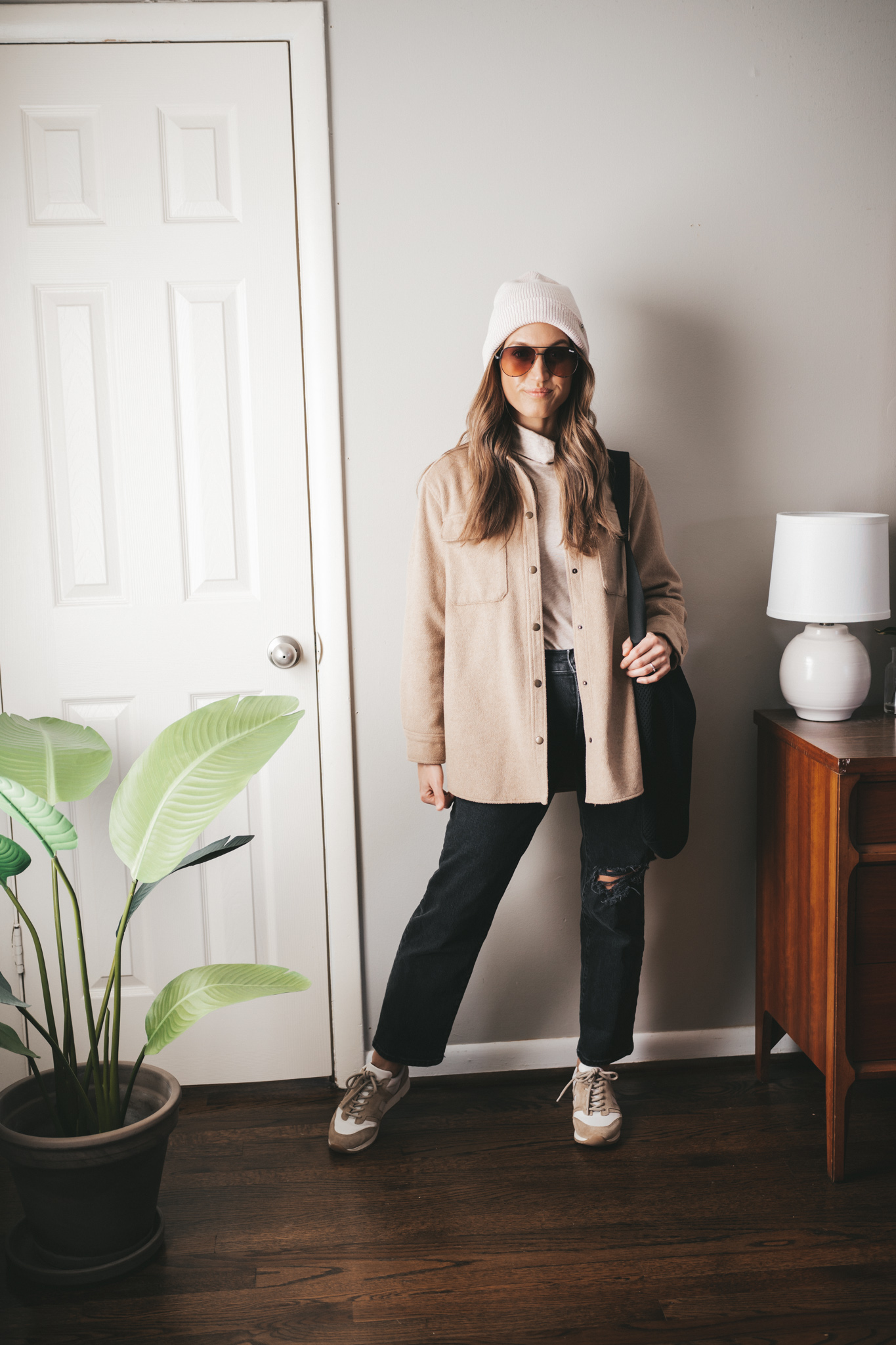 winter capsule wardrobe outfit with a turtleneck, tan shacket, black denim, white and tan sneakers, a cream colored beanie and oversized aviator sunglasses