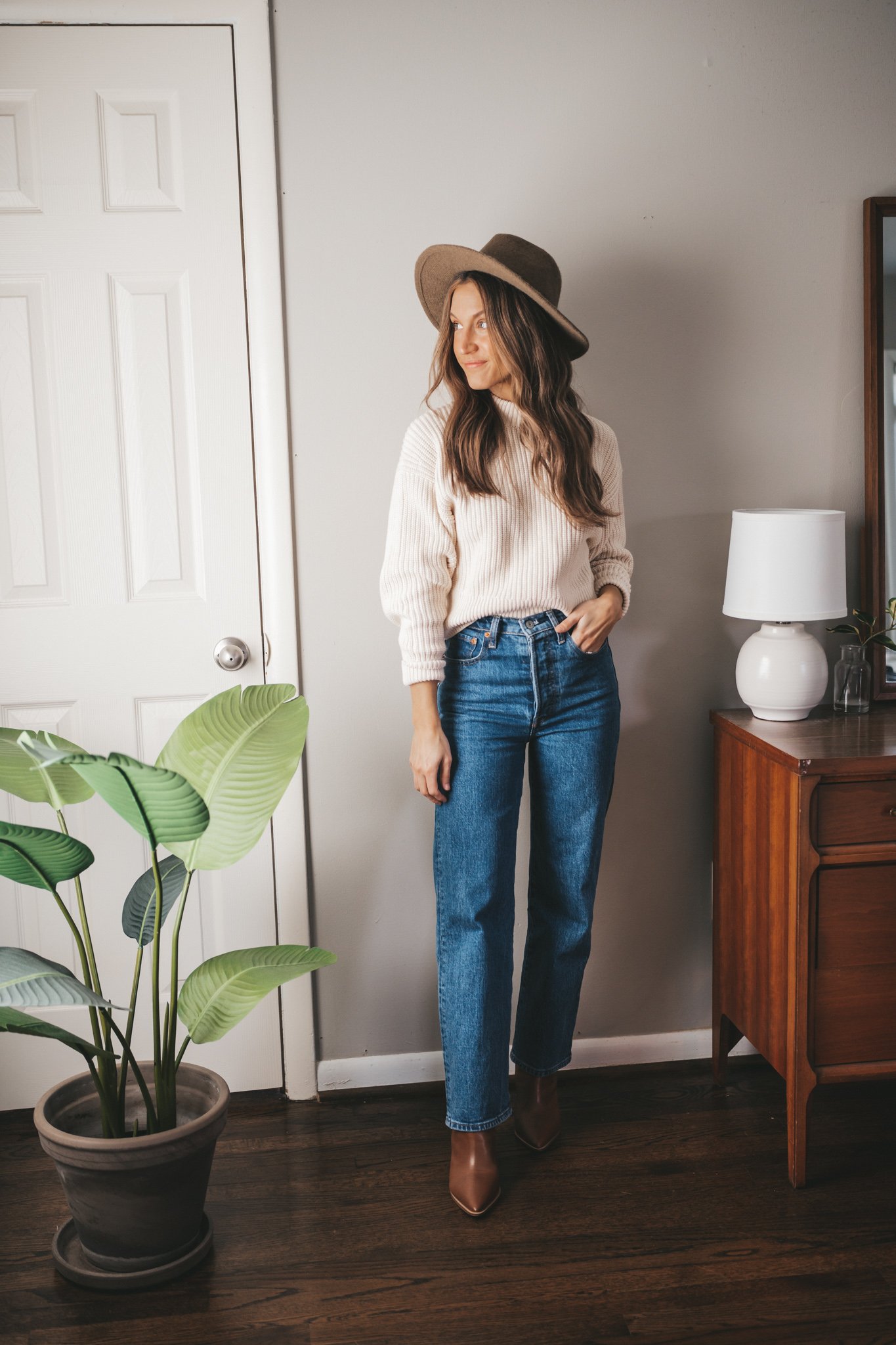 ashley wearing a cream colored chunky crewneck sweater, dark wash straight denim, brown ankle boots, and a taupe fedora
