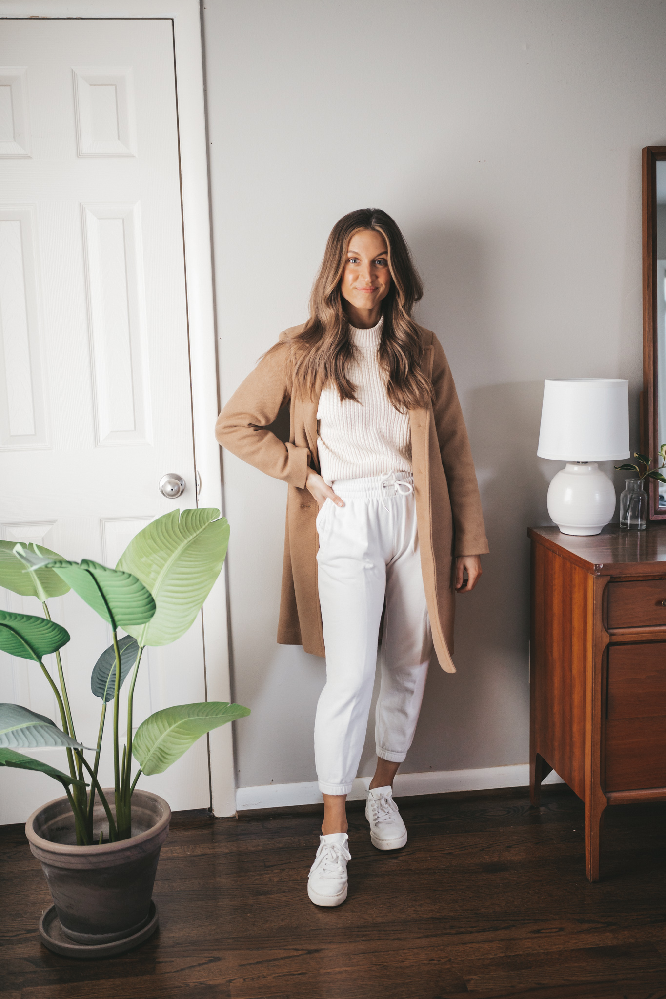 ashley wearing a cream colored crewneck sweater with white joggers, white sneakers, and a camel coat