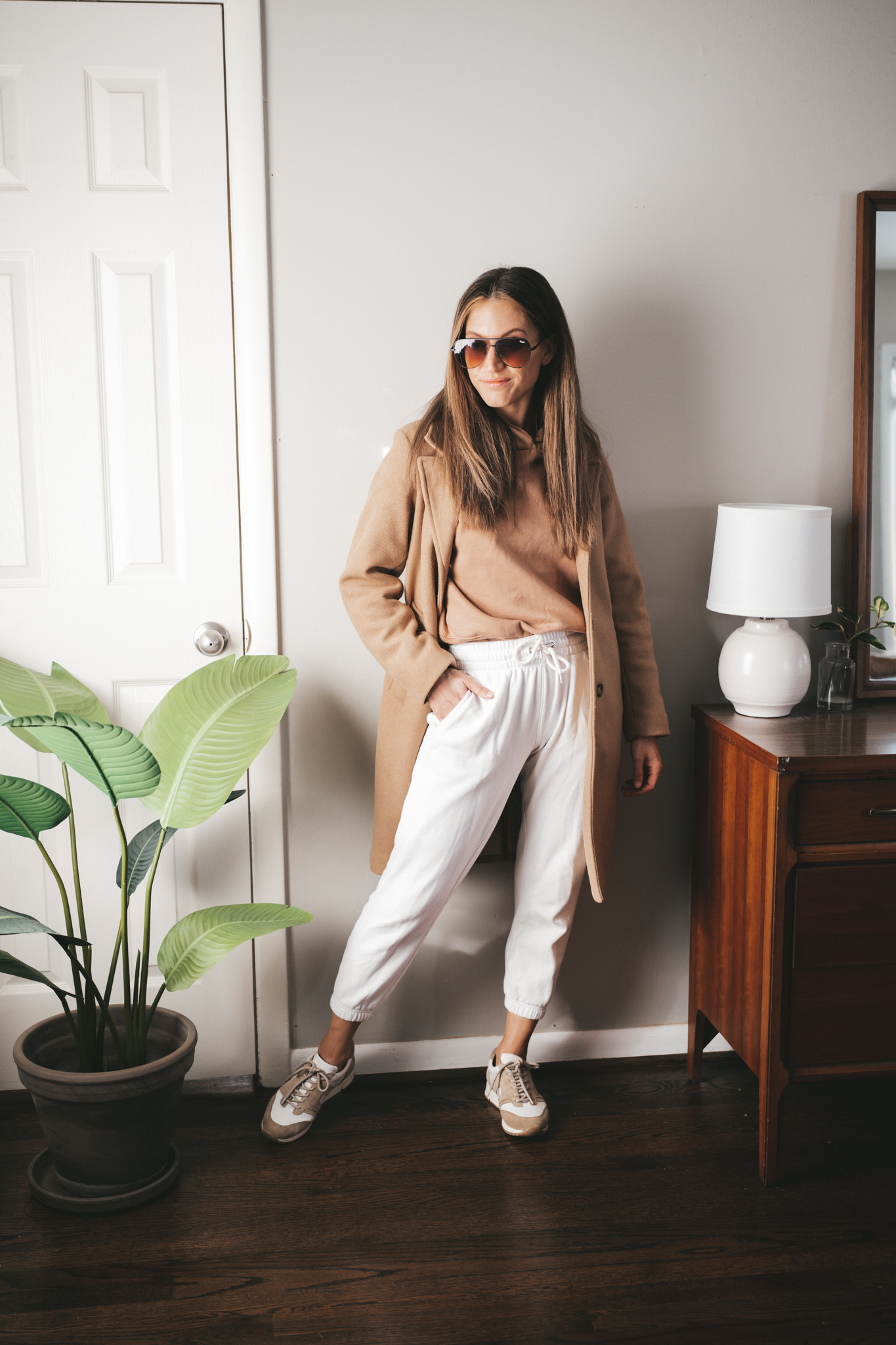 winter outfit idea with a tan hoodie, white joggers, camel coat, and white and tan leather sneakers with oversized aviator sunglasses