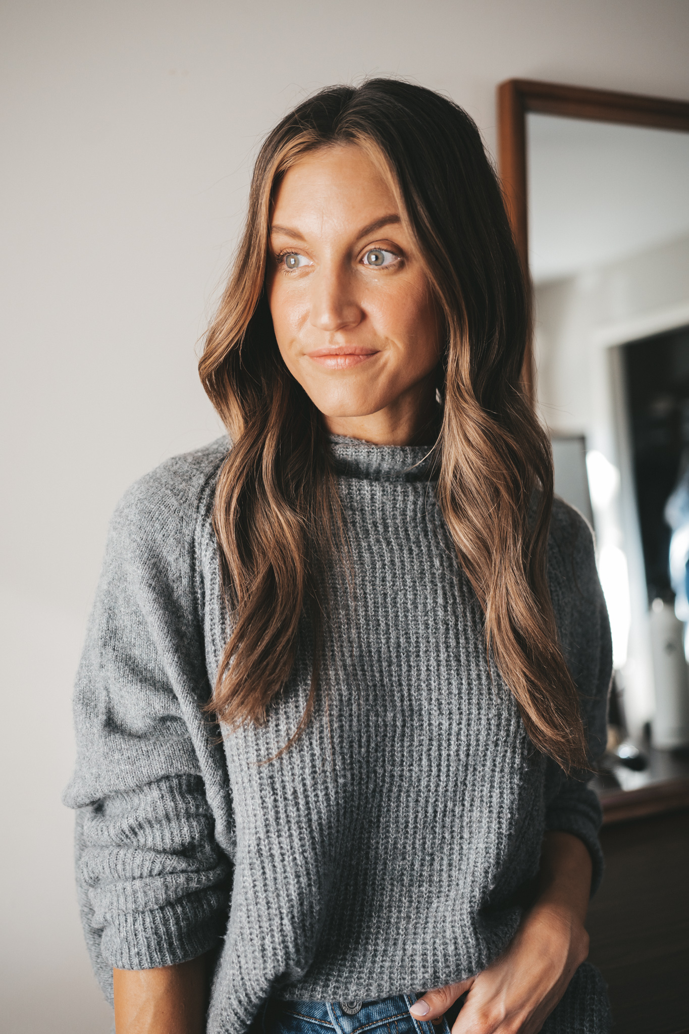 ashley with wavy curls standing in her bedroom in front of a mirror wearing a gray sweater