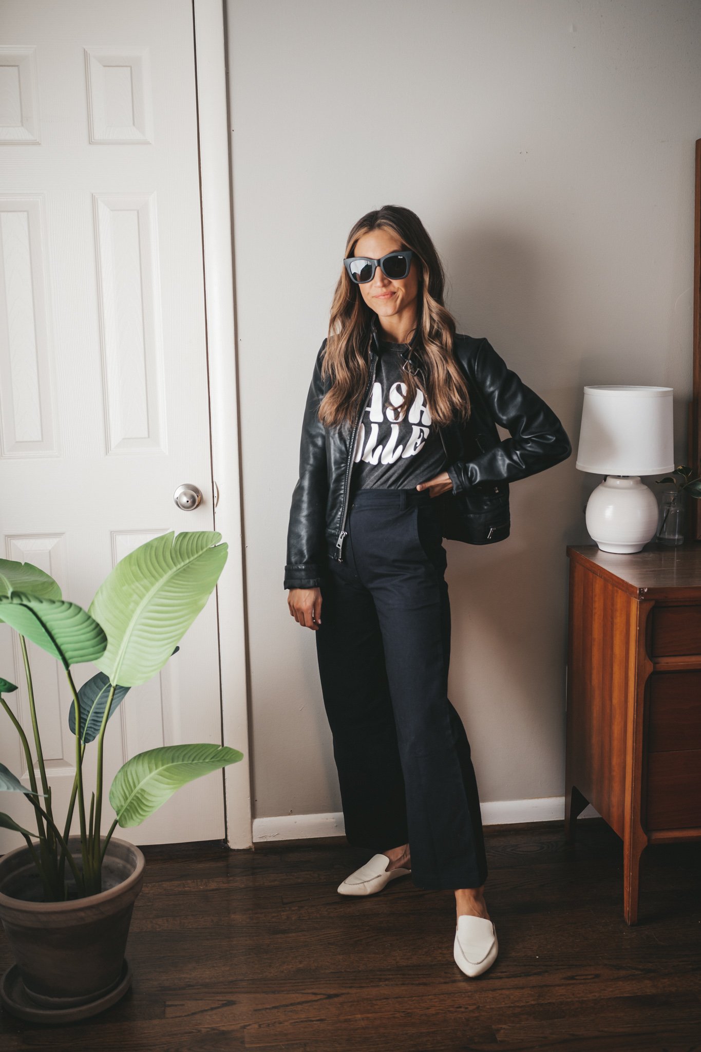 winter outfit with a black graphic tee, black faux leather moto jacket, black wide leg pants, white mules, and black square frame sunglasses