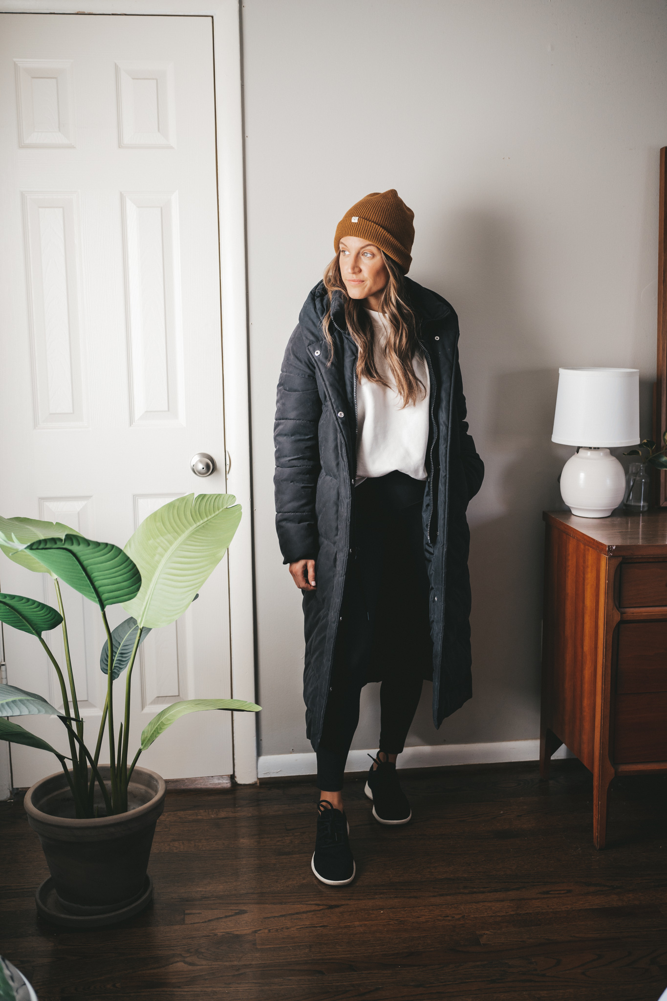 winter outfit idea with a cream colored sweatshirt, black leggings, black wool sneakers, black long puffer coat, and brown beanie