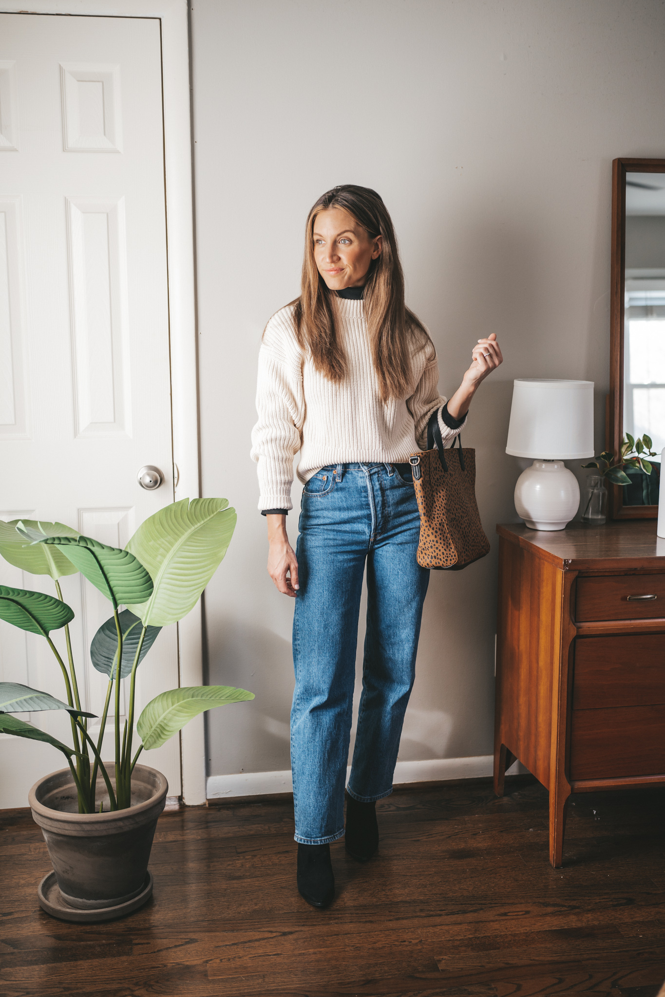 winter capsule wardrobe outfit with a cream colored sweater worn over a black turtleneck, mid-wash straight denim, black heeled ankle boots, and a leopard tote bag