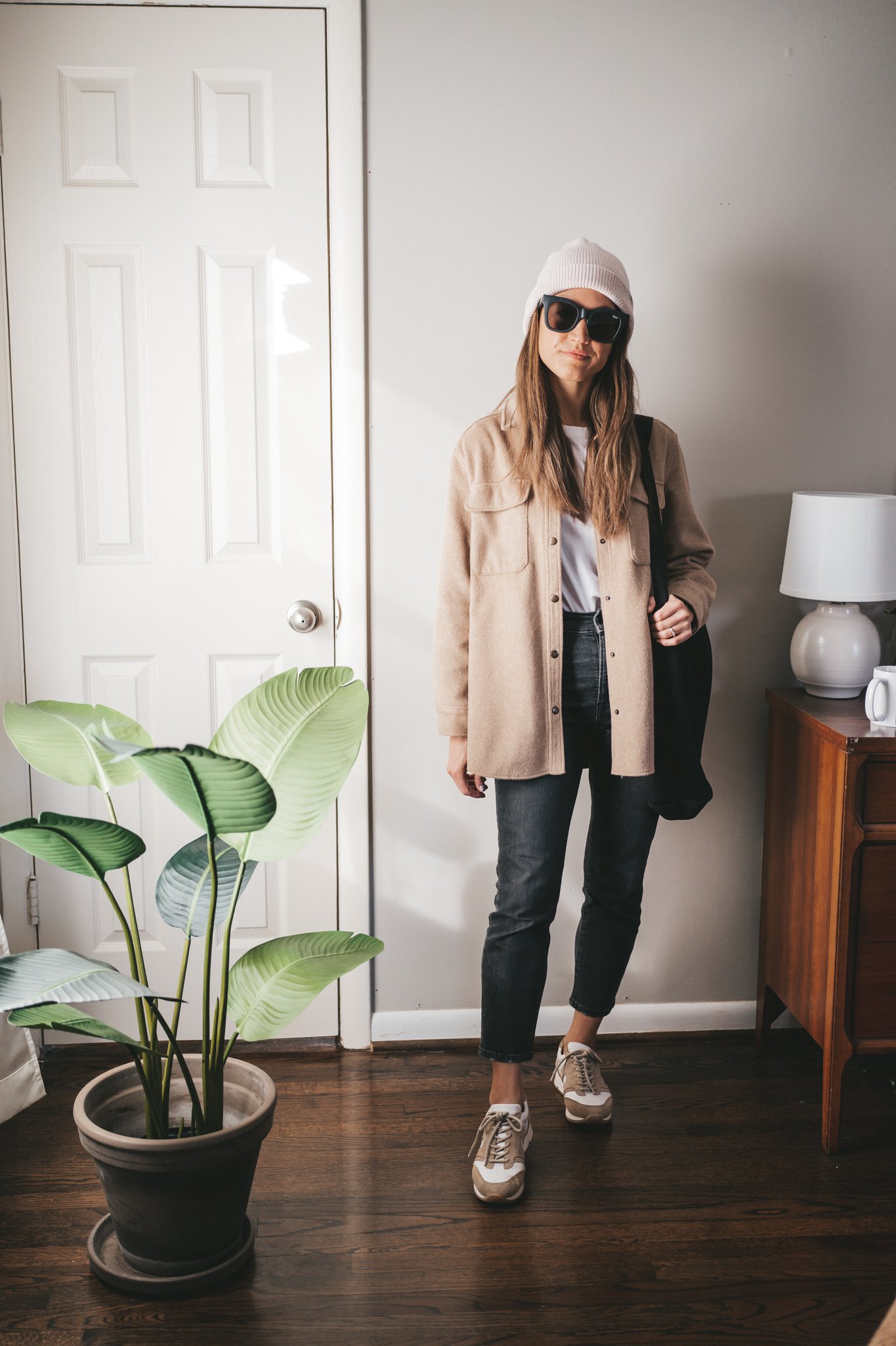 winter capsule wardrobe outfit with a white tee, tan shacket, washed black denim, white and tan leather sneakers, a cream colored beanie, and a black mesh tote bag