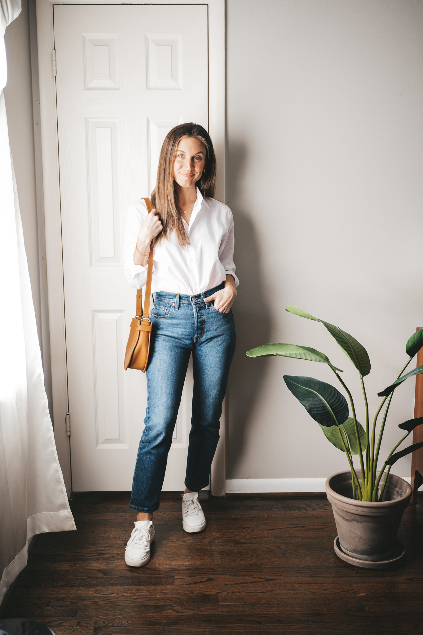 a winter mini capsule wardrobe outfit with a white button down, dark wash straight denim, white sneakers, and a cognac crossbody bag