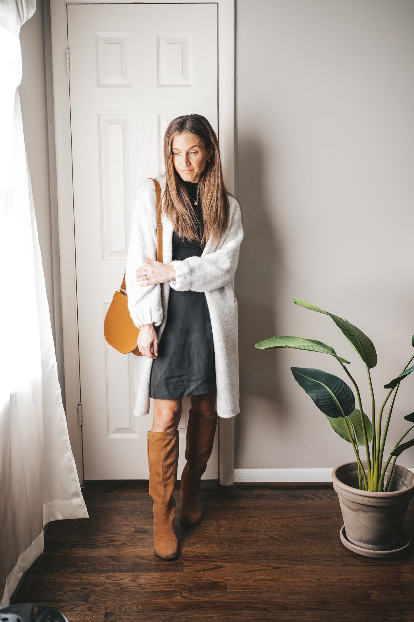 a winter mini capsule wardrobe outfit with a black polo tee, light gray sweater coat, black mini skirt, brown suede knee high boots, and a cognac crossbody bag