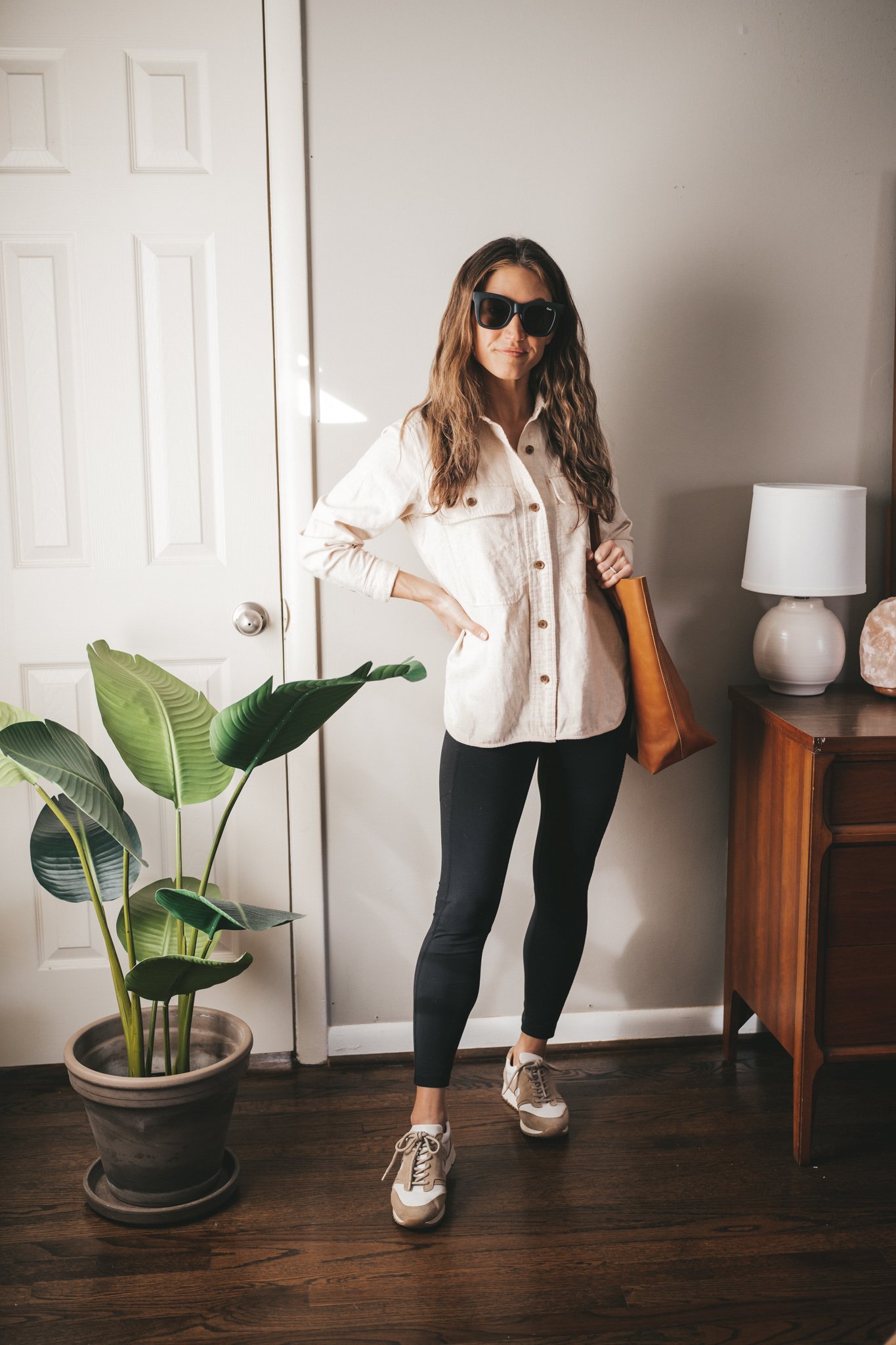 winter outfit with a cream colored flannel, black leggings, white and tan sneakers, a cognac leather tote bag and black square frame sunglasses