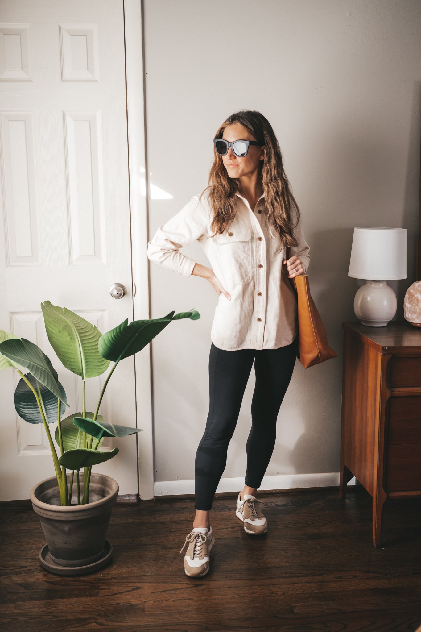 winter outfit with a cream colored flannel, black leggings, white and tan sneakers, a cognac leather tote bag and black square frame sunglasses