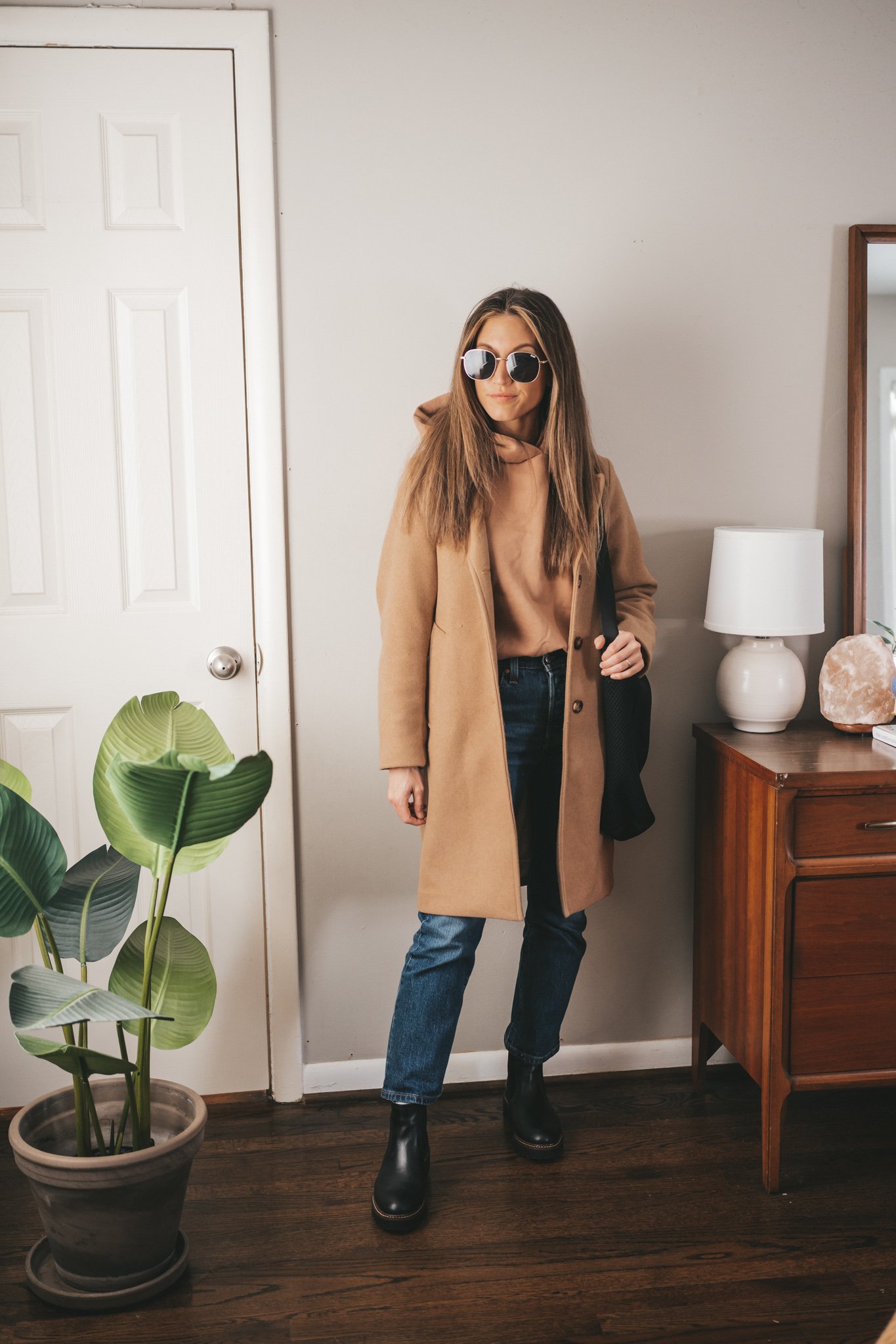 winter outfit with a tan hoodie, camel coat, mid-wash straight denim, and black lug sole chelsea boots worn with a black tote bag, and gold frame sunglasses