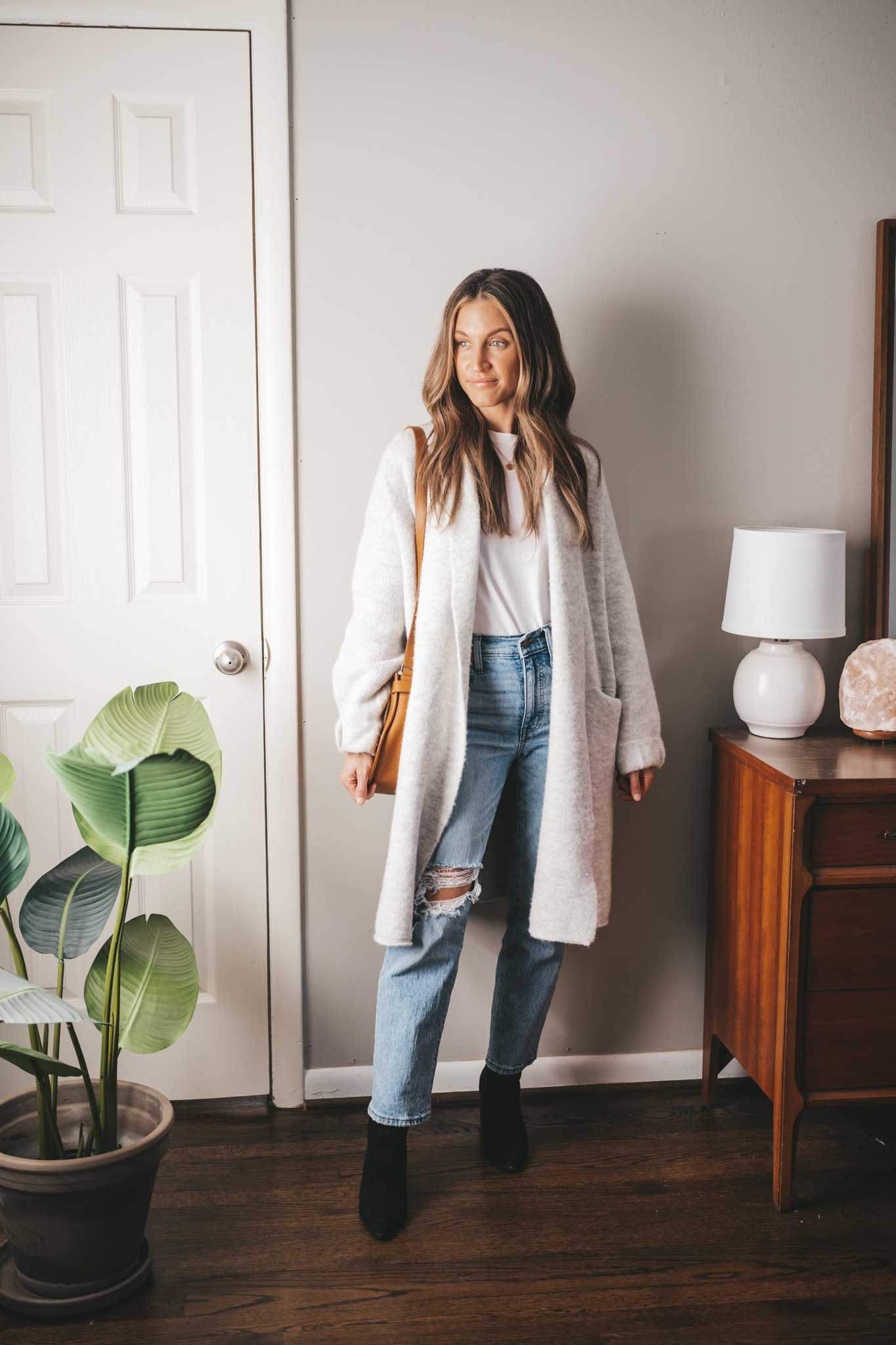 winter outfit with a white tee, light gray sweater coat, light wash distressed denim, black heeled ankle boots, and a cognac crossbody bag