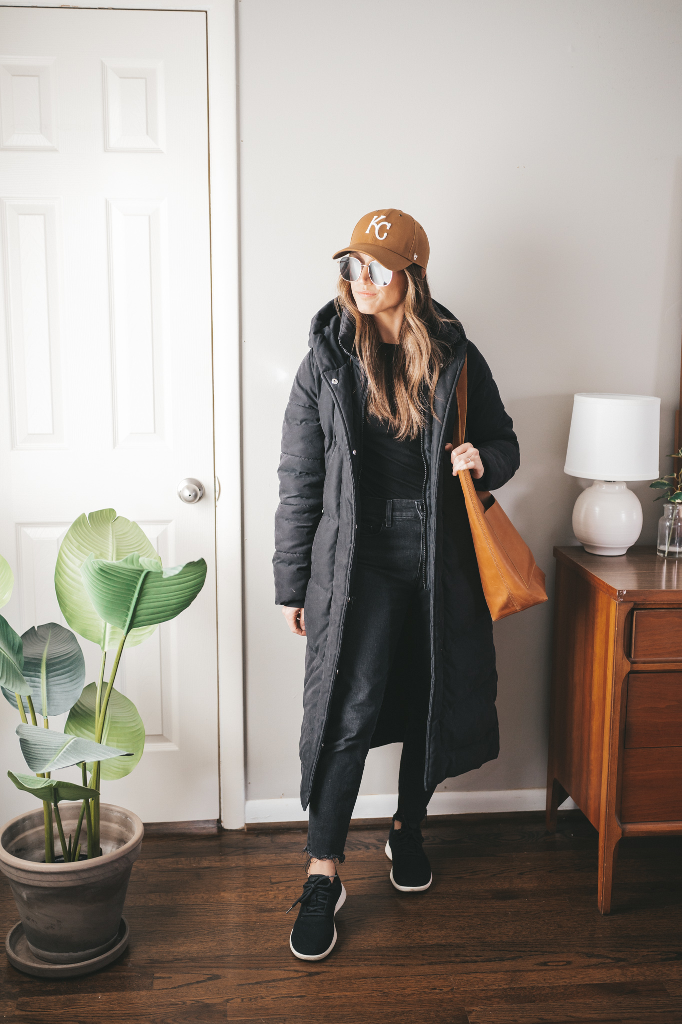 late winter outfit idea with a black tee, black denim, black wool sneakers, a black puffer coat, and a tan KC baseball hat with a cognac leather tote bag