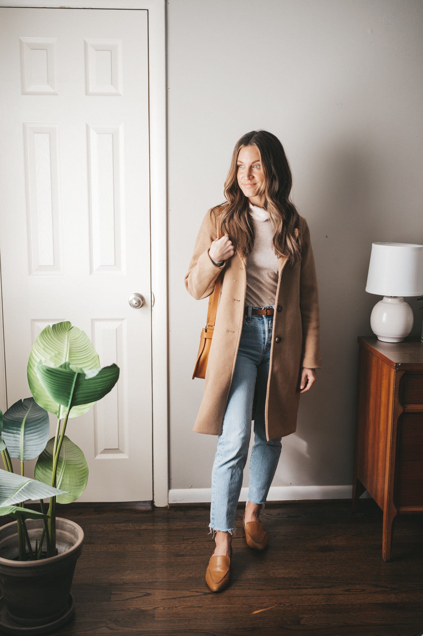 ashley wearing an ivory turtleneck, camel coat, light wash denim, cognac loafers, and a cognac crossbody bag