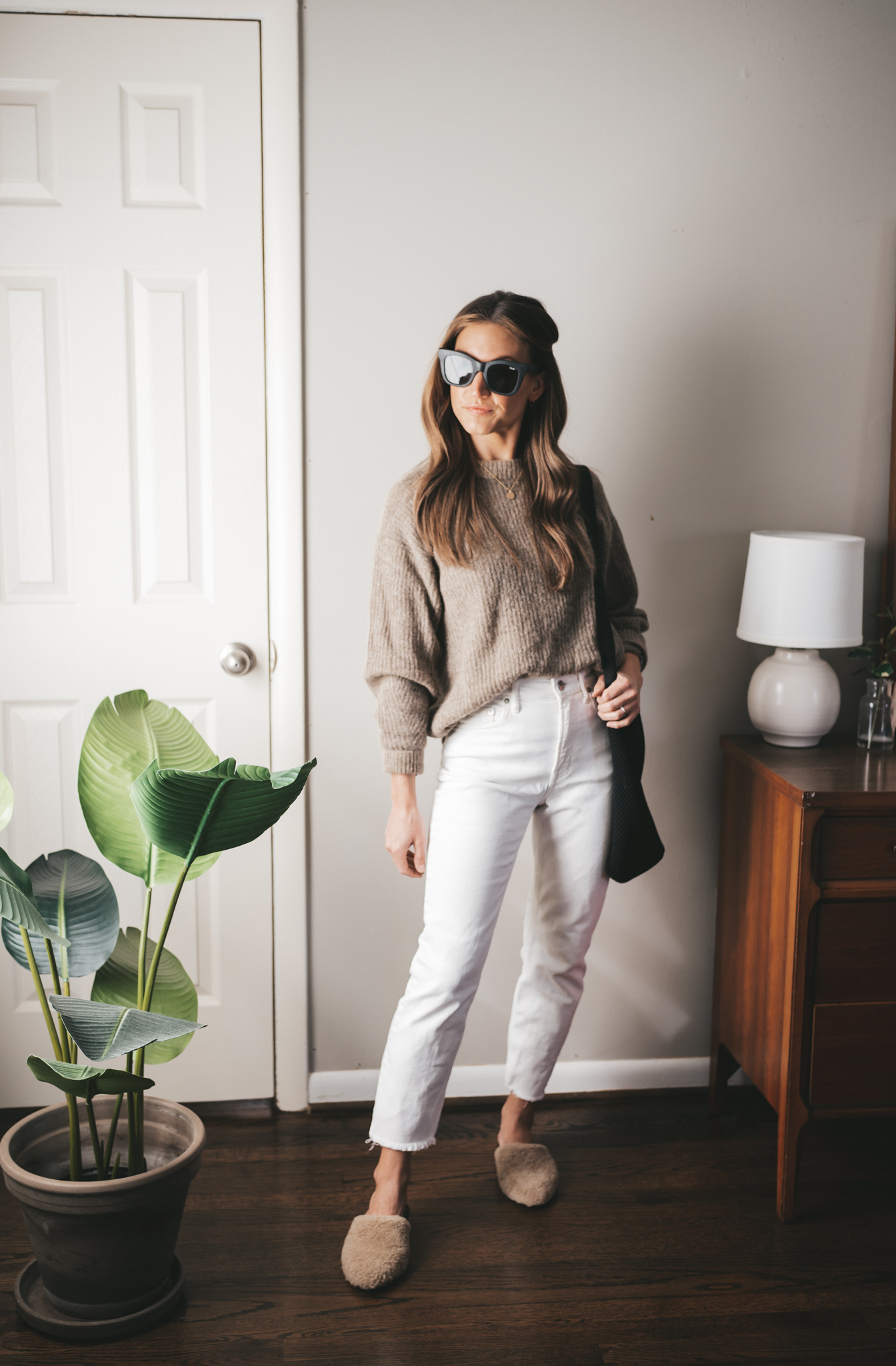 late winter outfit idea with a taupe alpaca sweater, white straight denim, shearling mules, a black tote bag, and black square frame sunglasses