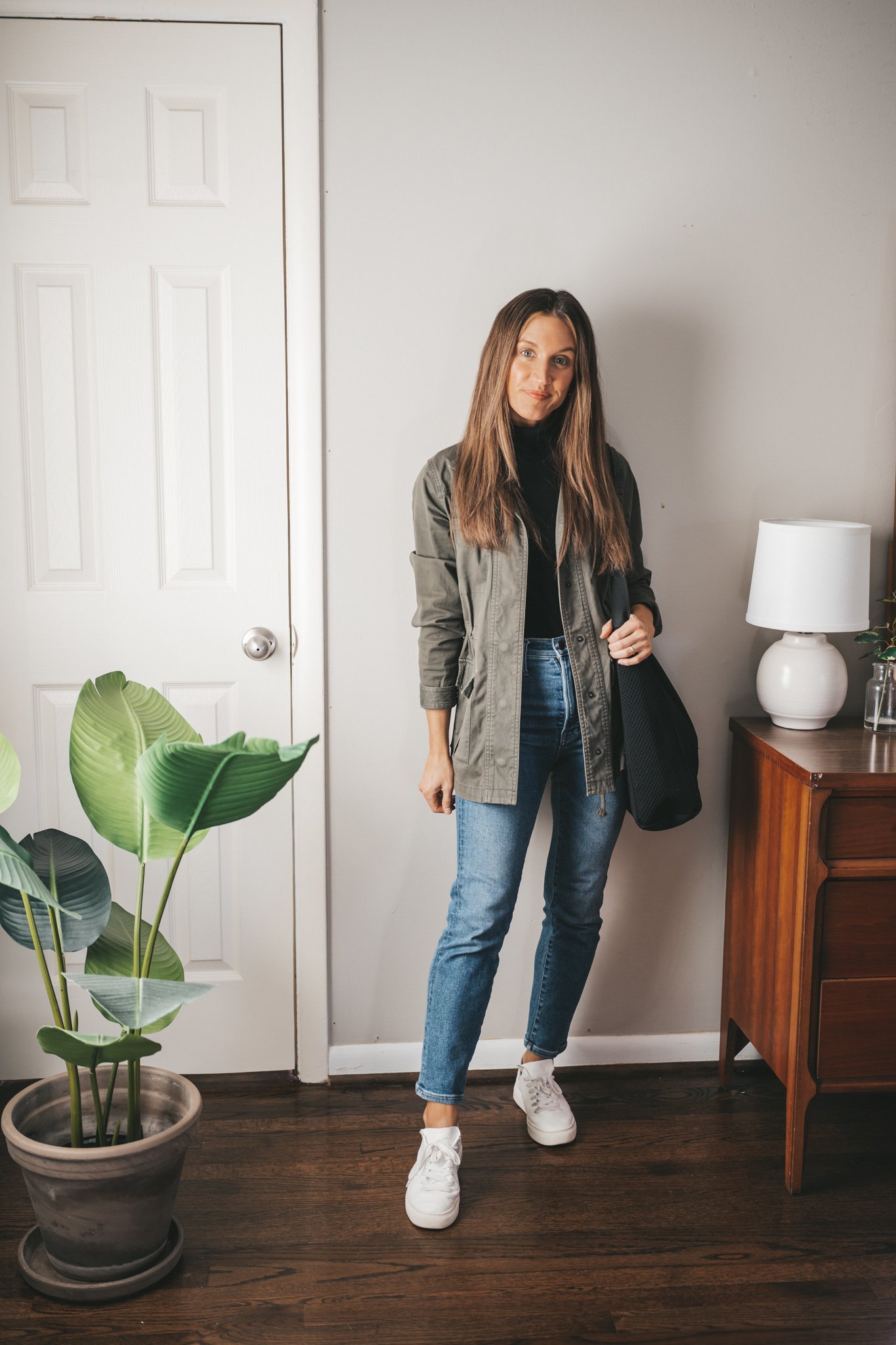 ashley wearing a black turtleneck outfit with a green utility jacket, dark wash mom jeans, white sneakers, and a black mesh tote bag