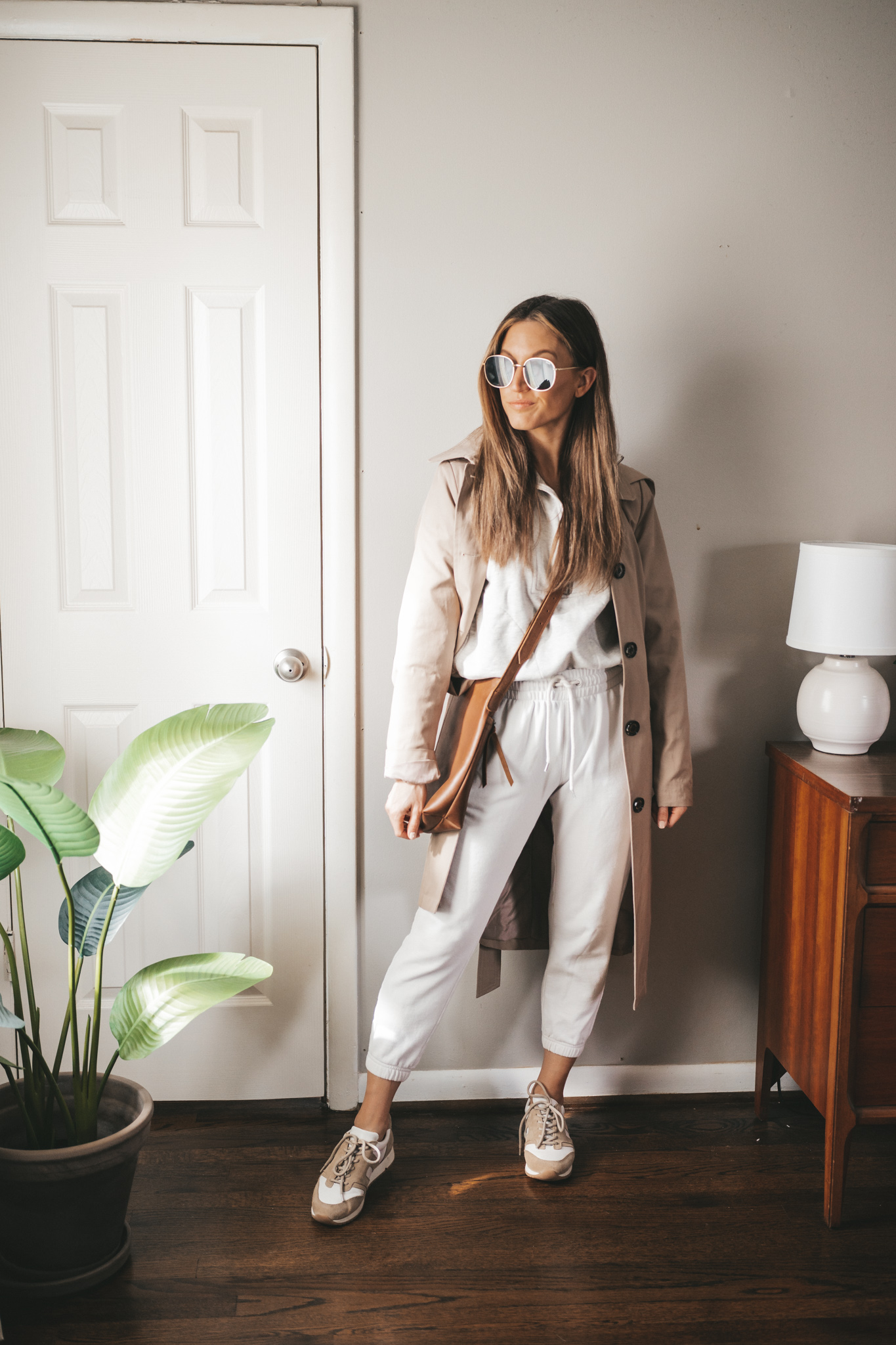 spring outfit idea with a light gray half zip sweatshirt, white joggers, white and tan sneakers, taupe trench coat, and brown crossbody bag