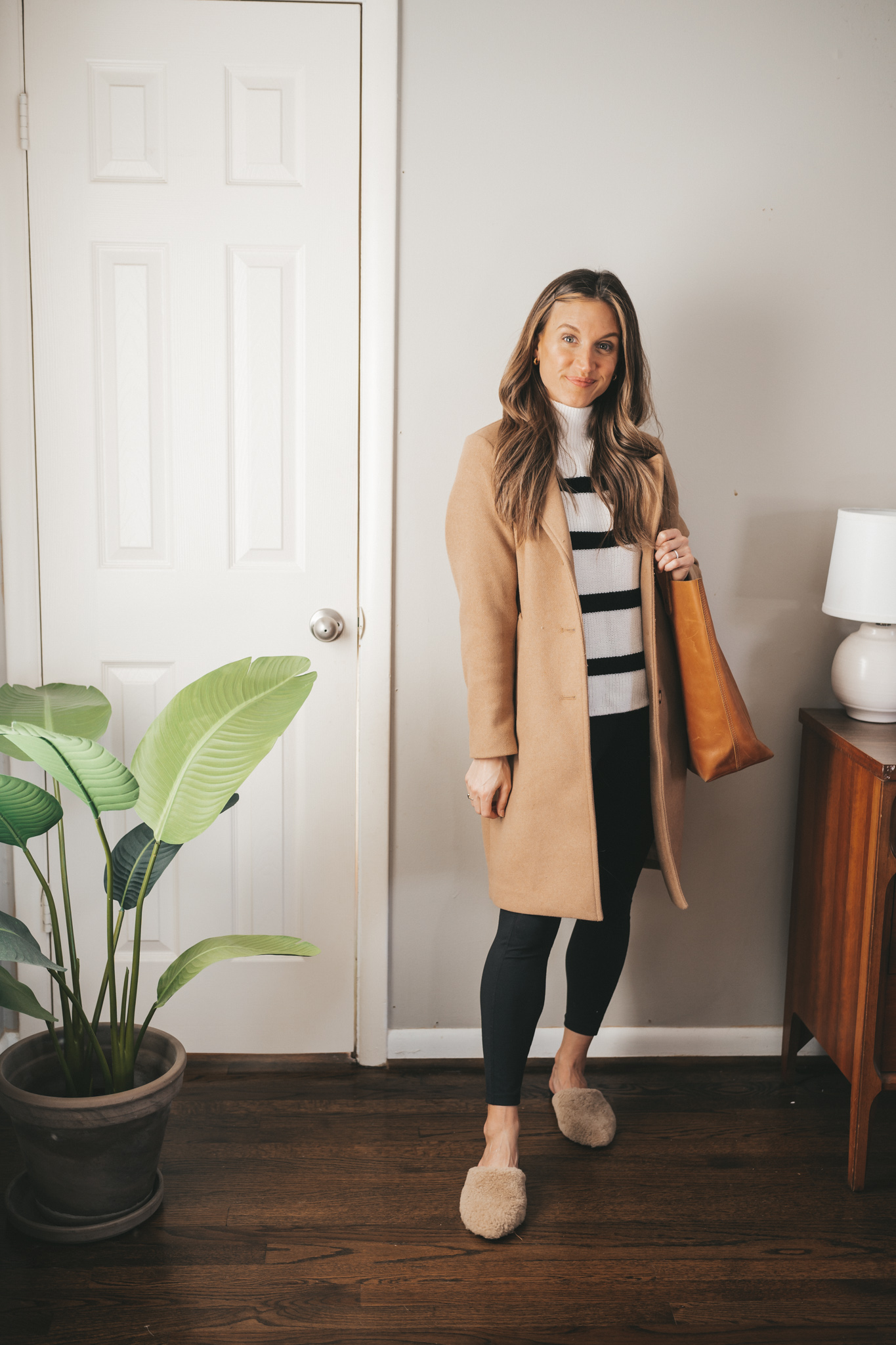 work from home outfit with a striped sweater, camel coat, black leggings, and shearling mules