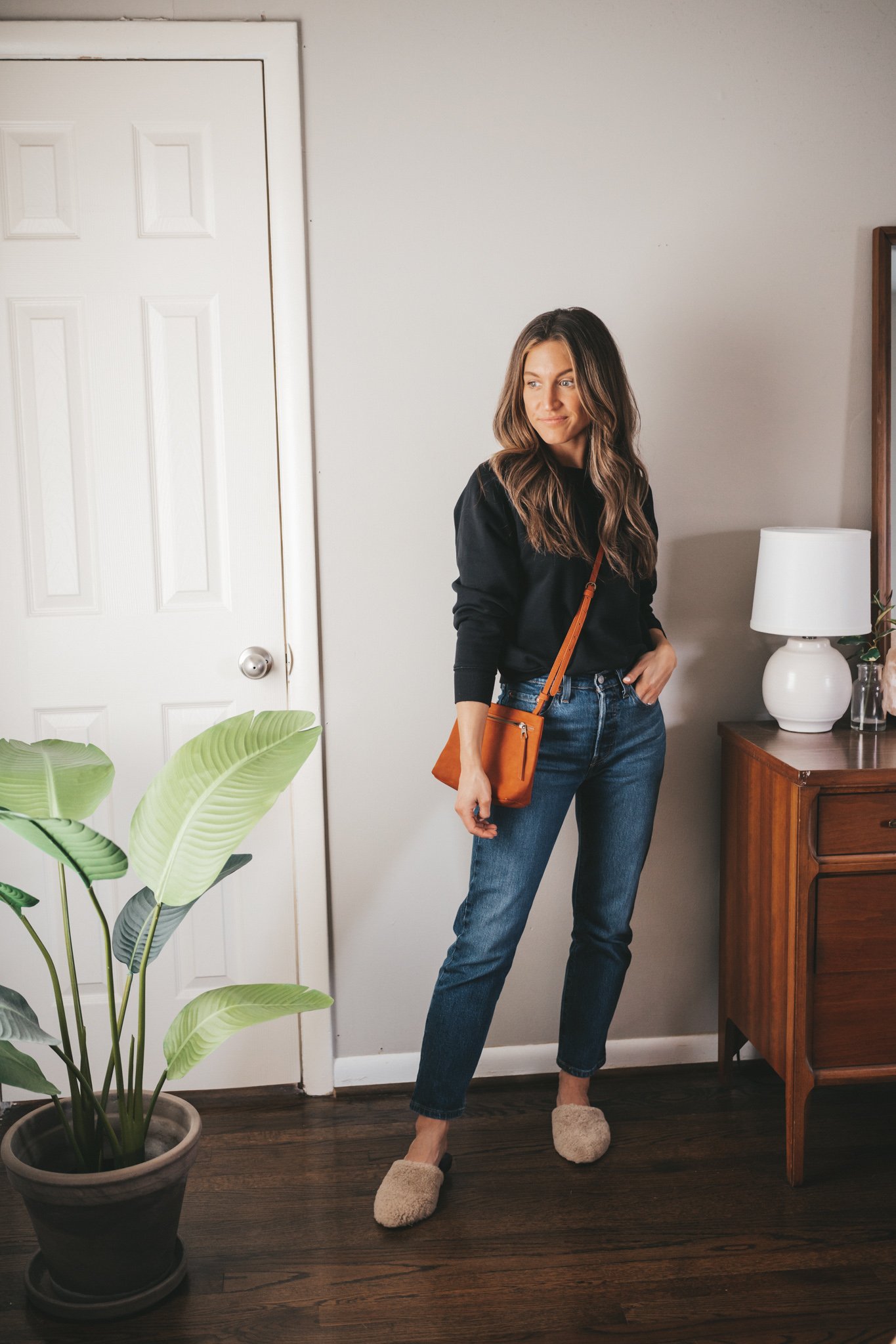 casual spring outfit idea with a black crewneck sweatshirt, dark wash straight denim, tan shearling mules and a red-orange crossbody bag