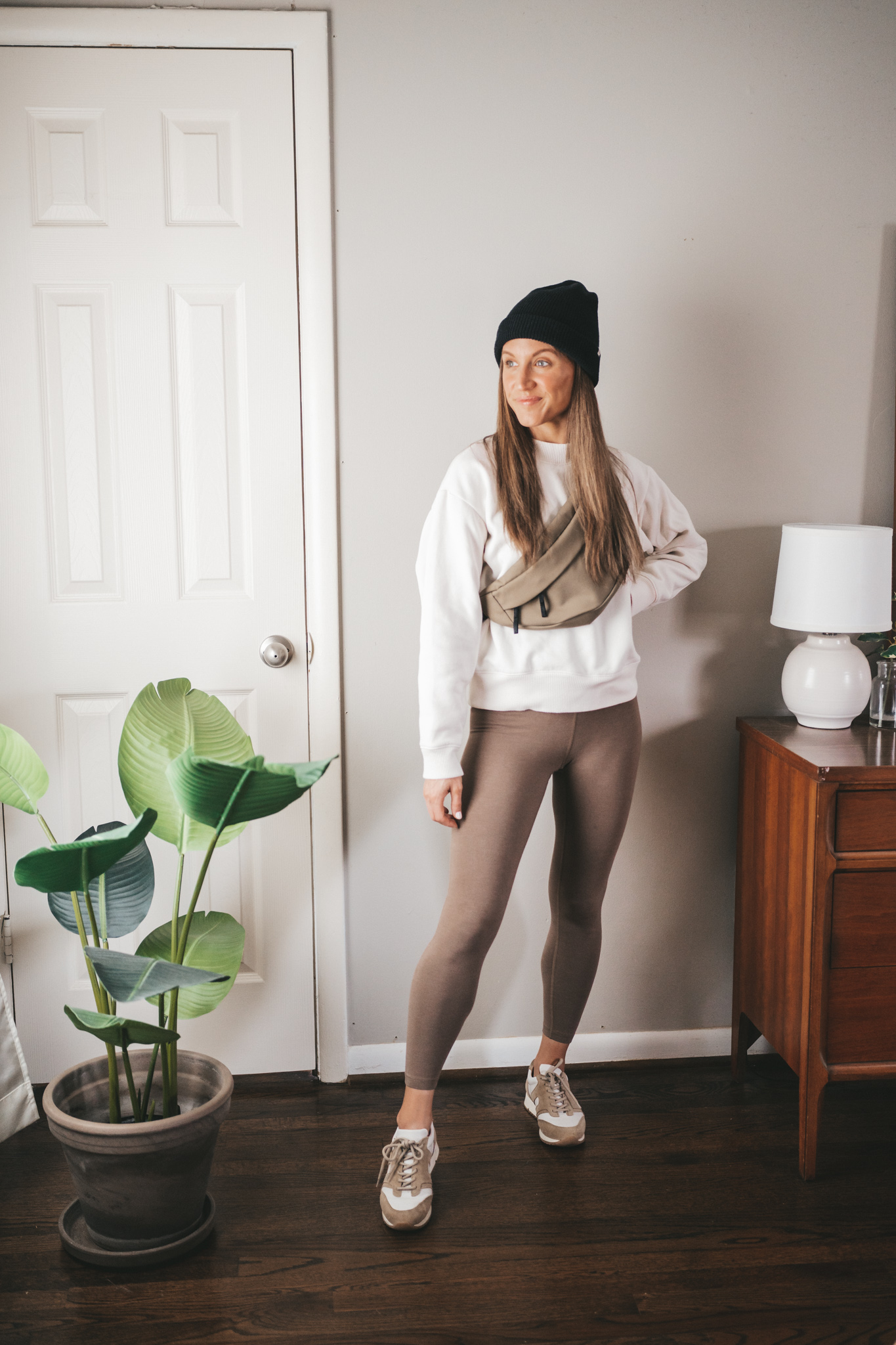casual spring outfit idea with taupe leggings, cream crewneck sweatshirt, white and tan sneakers, an olive green belt bag, and a black beanie