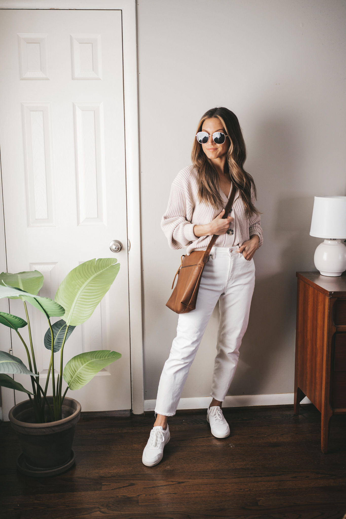 casual spring outfit ideas with a taupe button front sweater, white denim, white sneakers, a brown crossbody bag, and oversized gold frame sunglasses