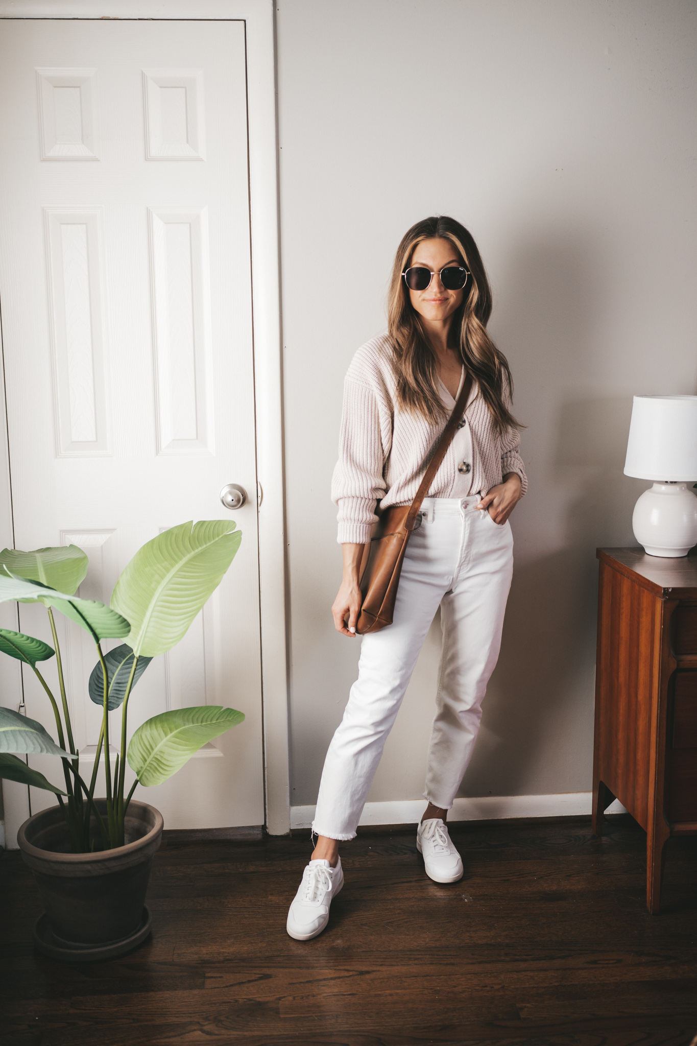 casual spring outfit ideas with a taupe button front sweater, white denim, white sneakers, a brown crossbody bag, and oversized gold frame sunglasses
