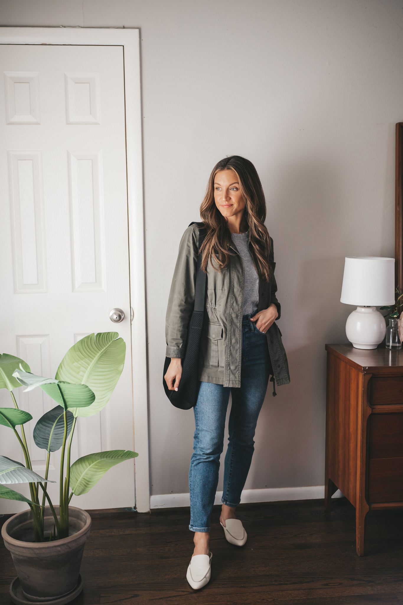 casual spring outfit idea with a gray tee, green utility jacket, mid-wash mom jeans, white mules, and a black tote bag
