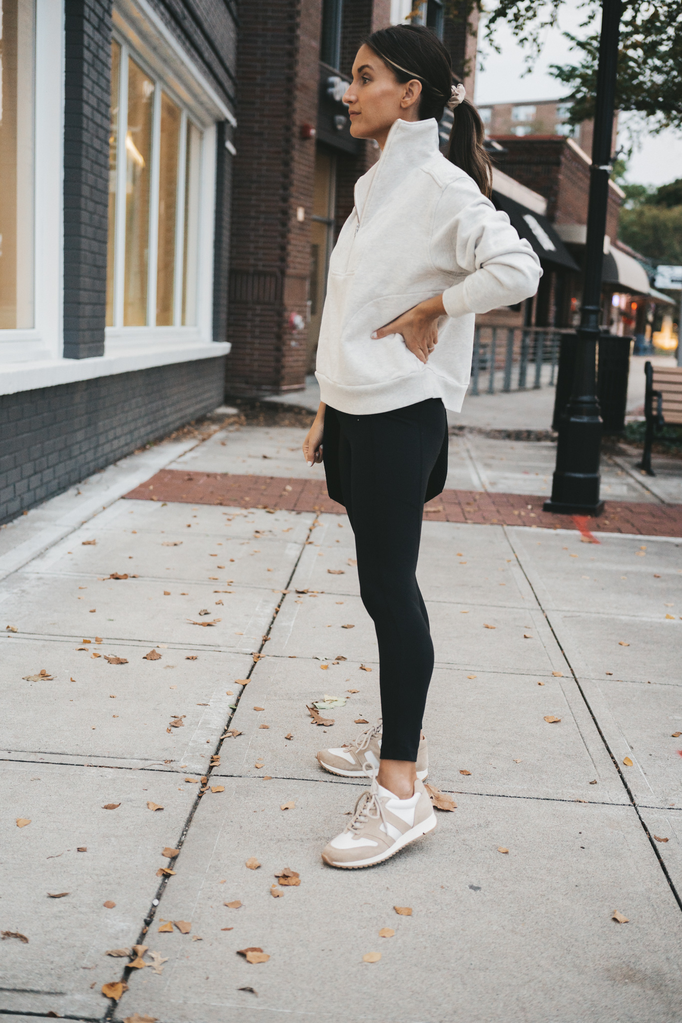 ashley wearing a gray half zip sweatshirt, black leggings, and tan and white leather sneakers standing on a sidewalk in front of a shop window