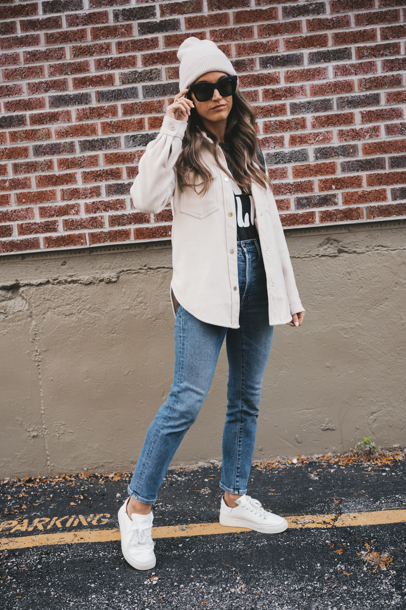 ashley wearing a washed black graphic tee that reads "Nashville", an ivory shacket, mid-wash denim, white sneakers, and a cream colored beanie standing in front of a brick wall