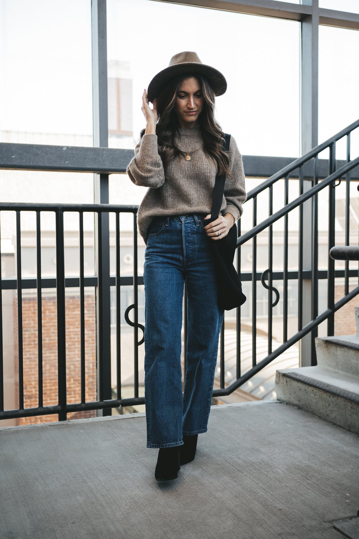 ashley wearing a taupe sweater, dark wash straight denim, black ankle boots, and a taupe fedora standing in an outdoor stairwell