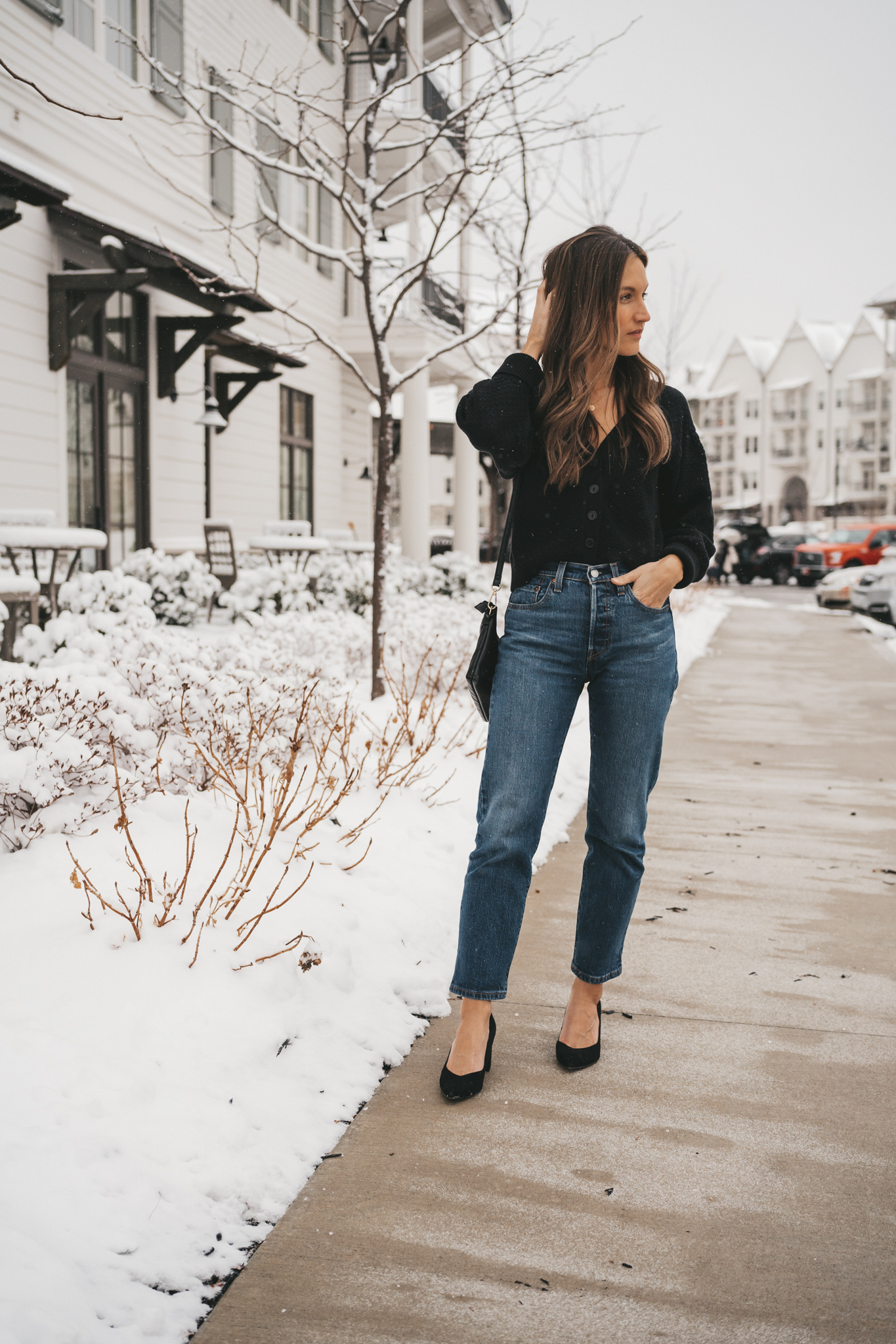 ashley wearing a cropped black buttoned cardigan, dark wash denim, and black pumps standing on a sidewalk in front of a white building with snow on the ground