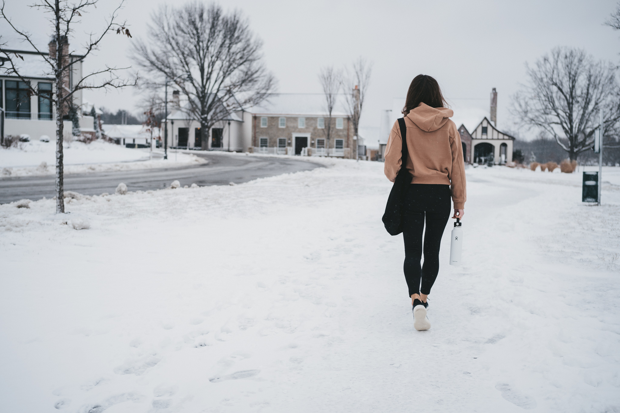 ashley wearing a tan hoodie, black leggings, and white sneakers walking through snow