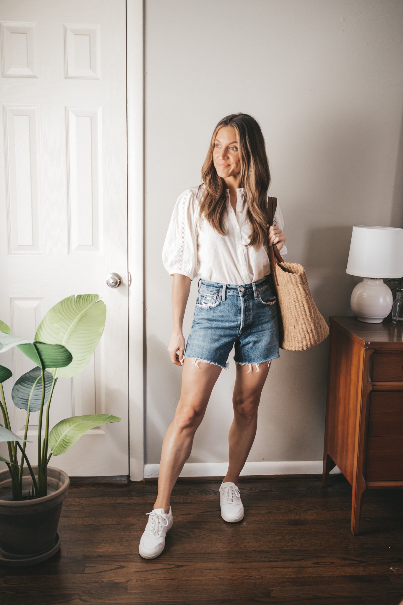 spring break outfit with a white blouse, denim cut-off shorts, white sneakers, and a straw tote bag