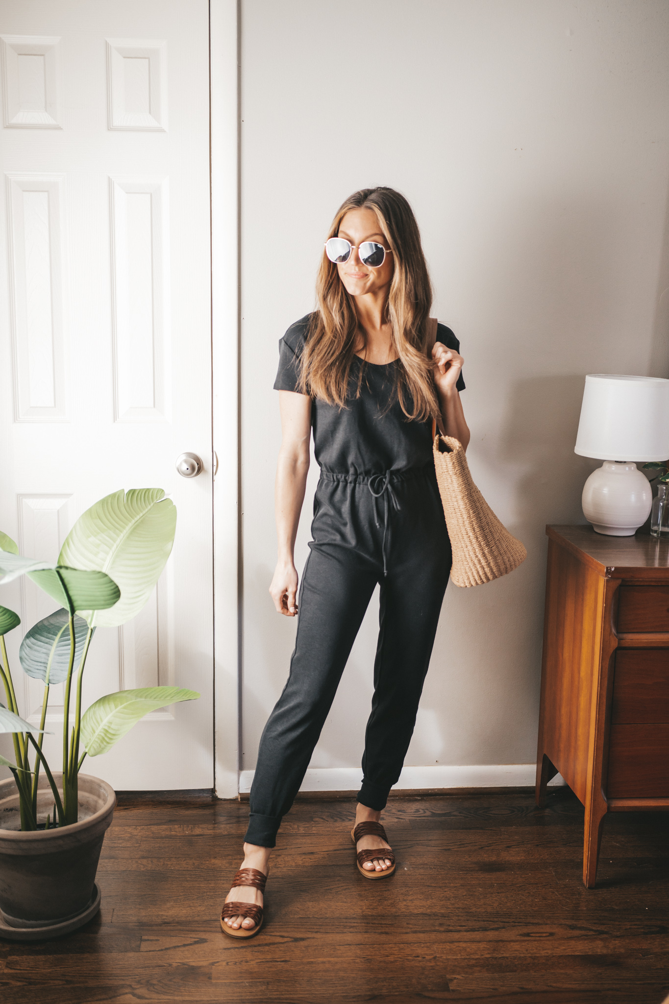 spring break outfit with a black drawstring jumpsuit, braided sandals, and a straw tote bag