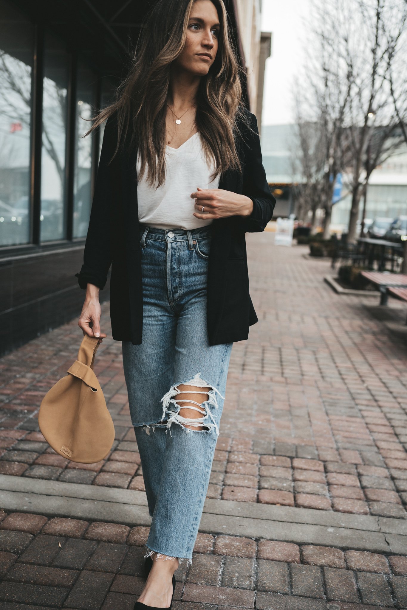 ashley wearing the agolde 90s pinch waist denim which a silk cami, black blazer, and black block heel pumps with a tan leather clutch bag walking down a paved sidewalk in front of some shops