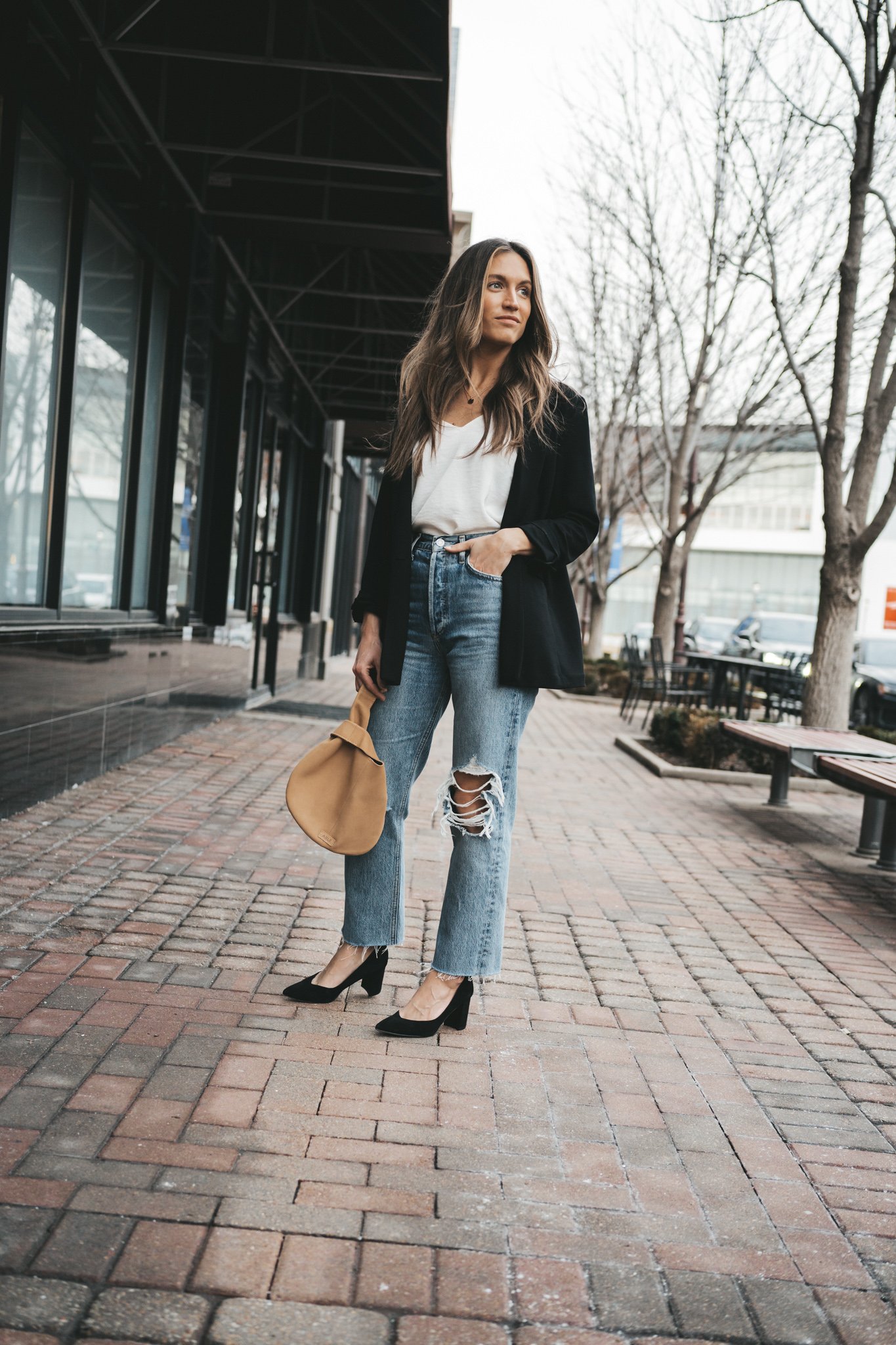 ashley wearing the agolde 90s pinch waist denim which a silk cami, black blazer, and black block heel pumps with a tan leather clutch bag walking down a paved sidewalk in front of some shops