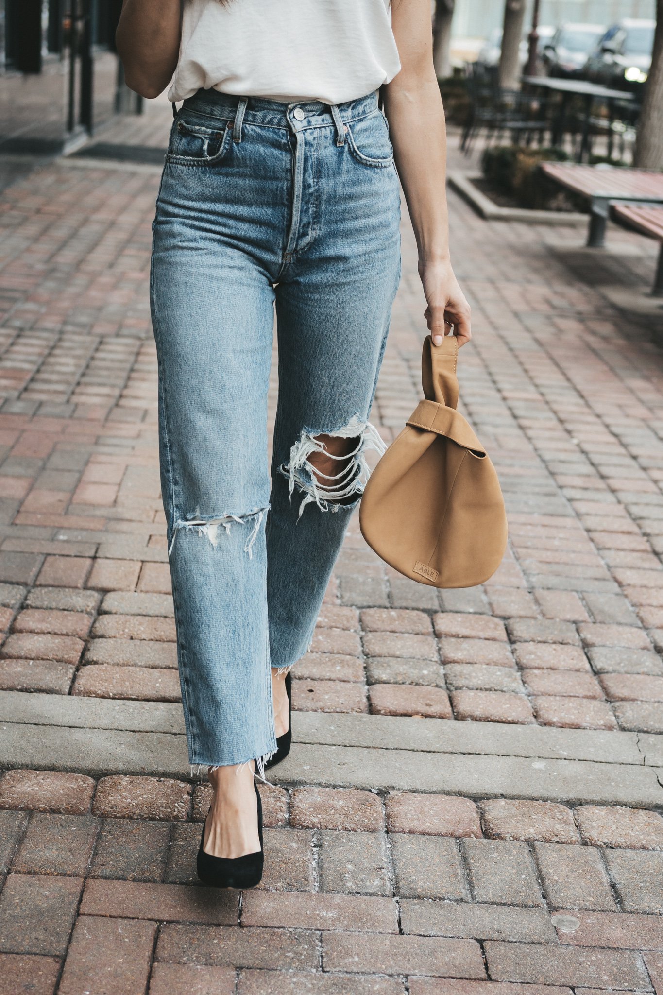 ashley wearing the agolde 90s pinch waist denim which a silk cami, black blazer, and black block heel pumps with a tan leather clutch bag walking down a paved sidewalk in front of some shops