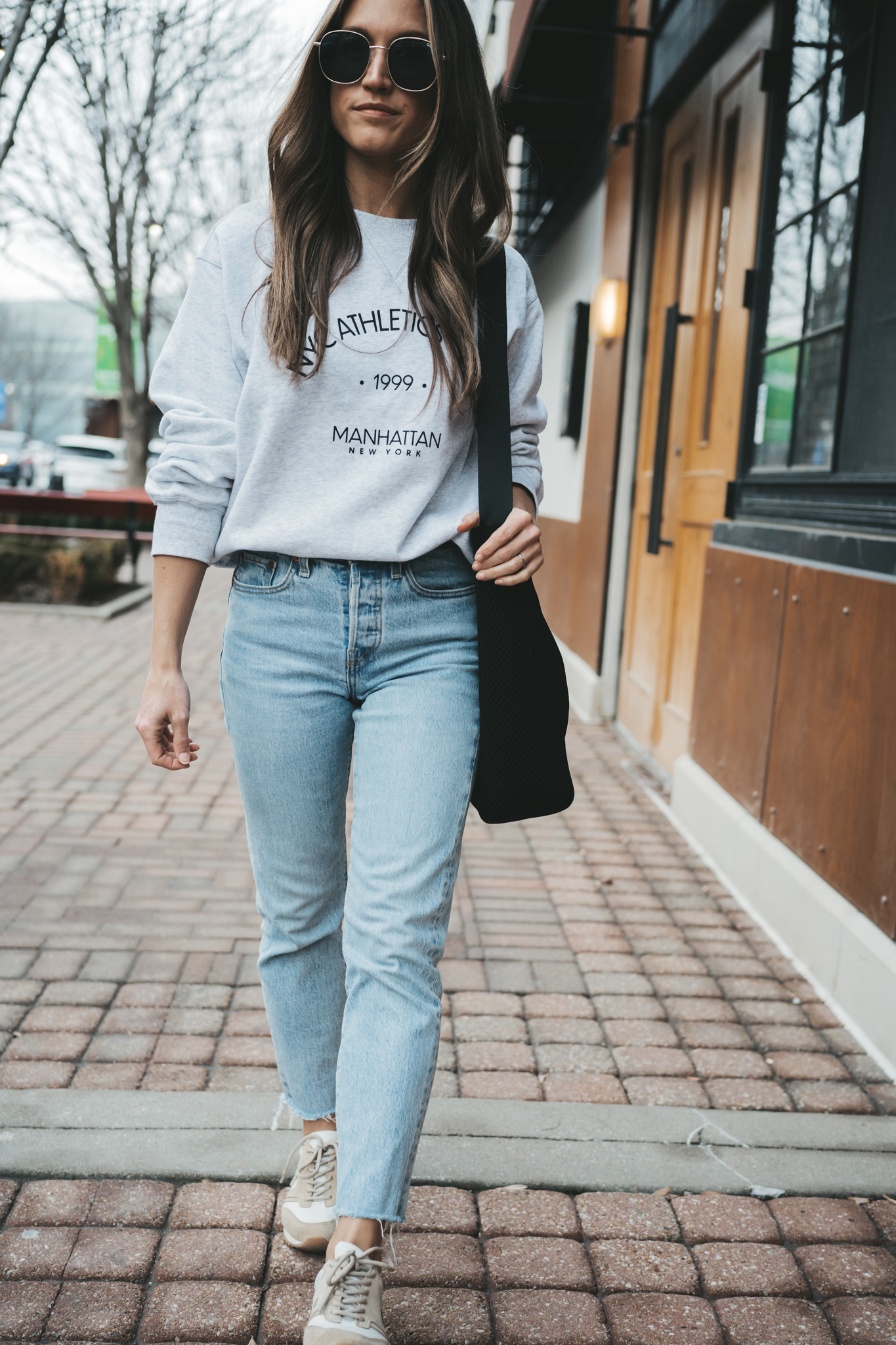 ashley wearing a gray graphic sweatshirt, light wash mom jeans, tan leather sneakers, oversized gold frame sunglasses and a black tote bag