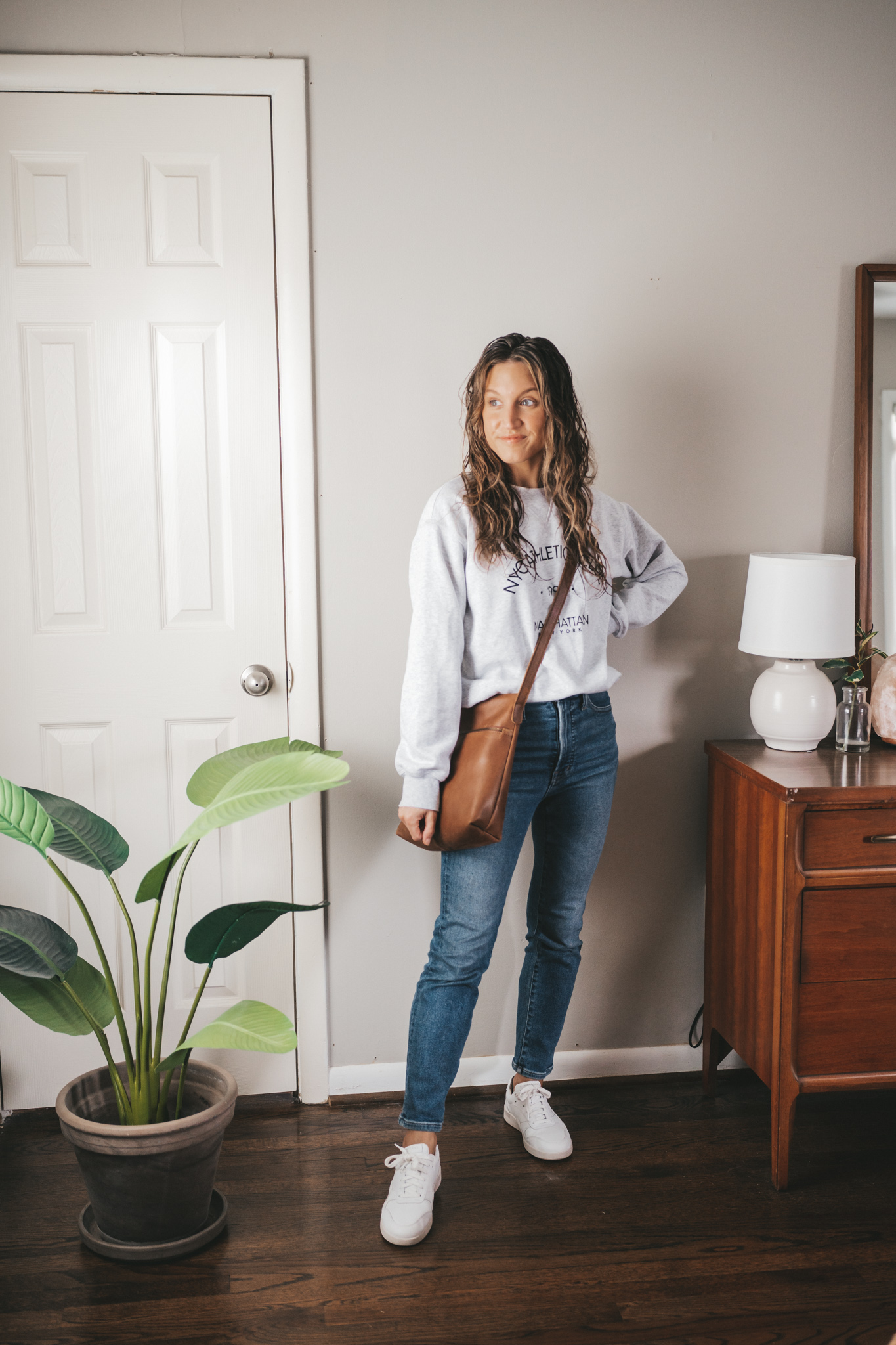 spring outfit with a gray graphic sweatshirt, mid-wash denim, white sneakers and a brown crossbody bag