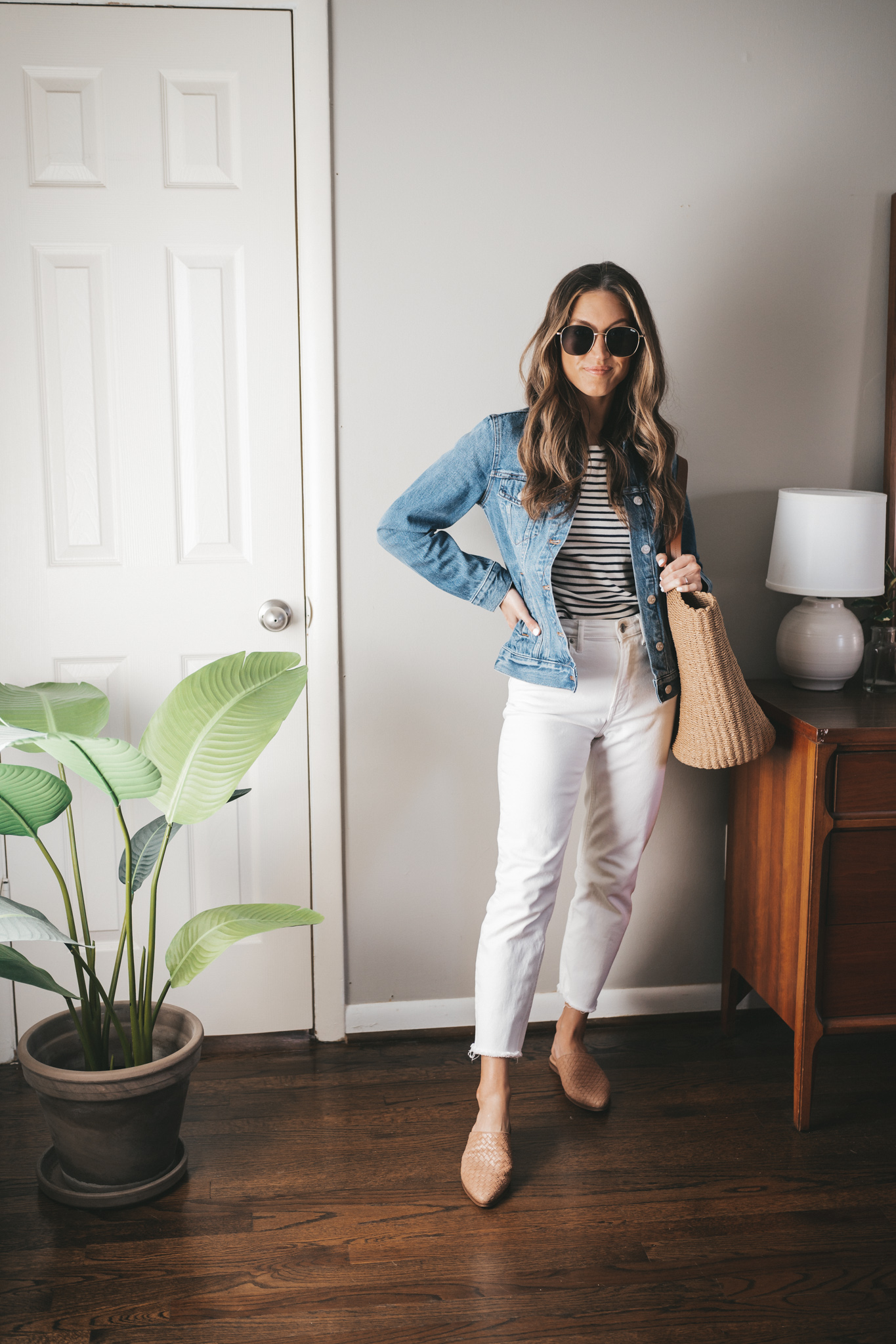 spring outfit with a blue and white striped tee, denim jacket, white denim, tan woven mules, and a straw tote bag