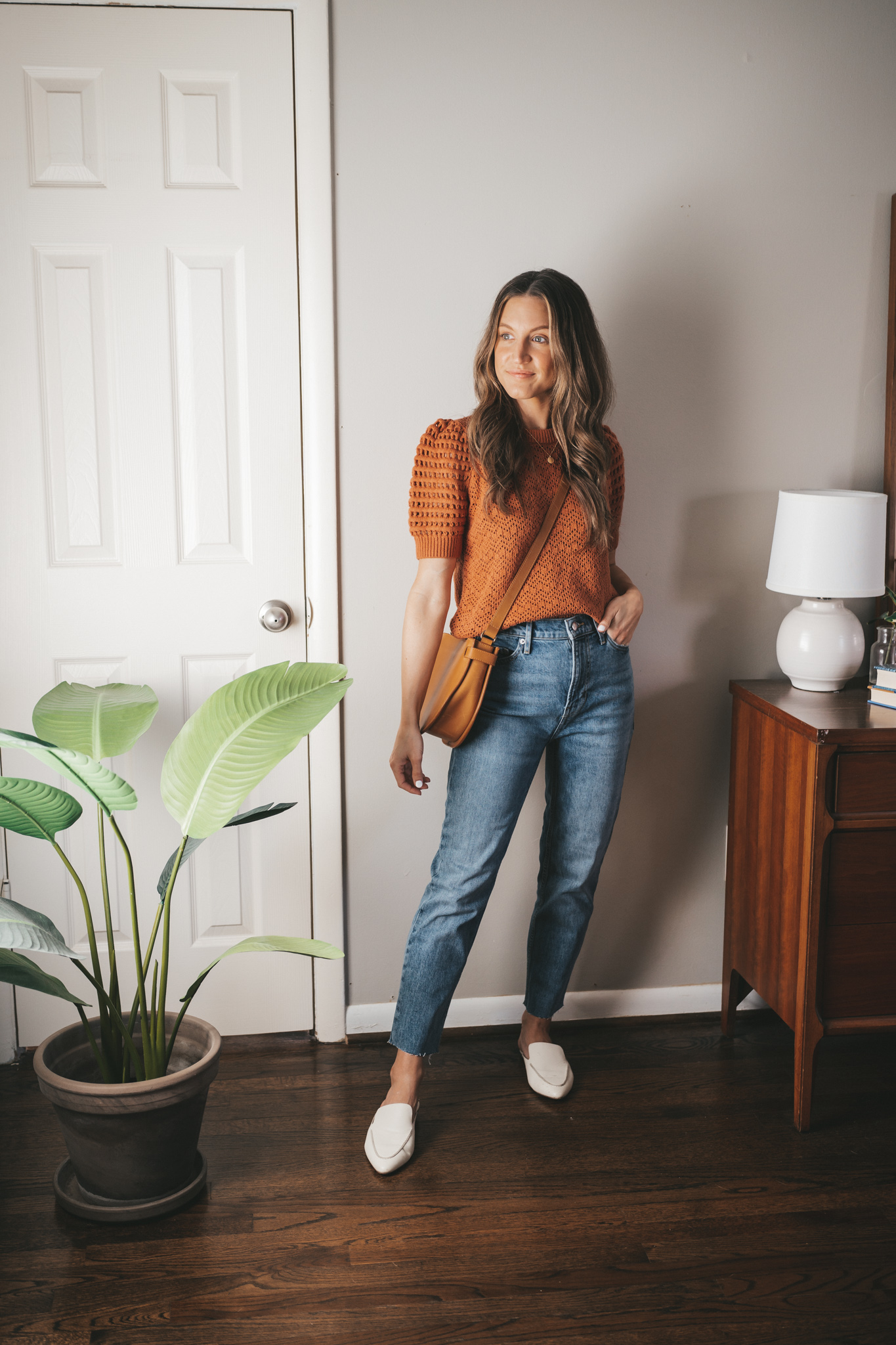 spring outfit with a rust colored knit sweater, mid-wash straight denim, white mules, and a cognac crossbody bag