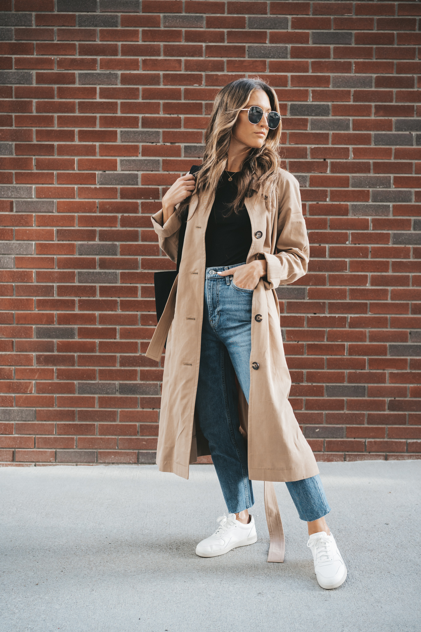ashley wearing a black tee, taupe trench coat, mid-wash denim, and white sneakers standing on a sidewalk in front of a brick wall