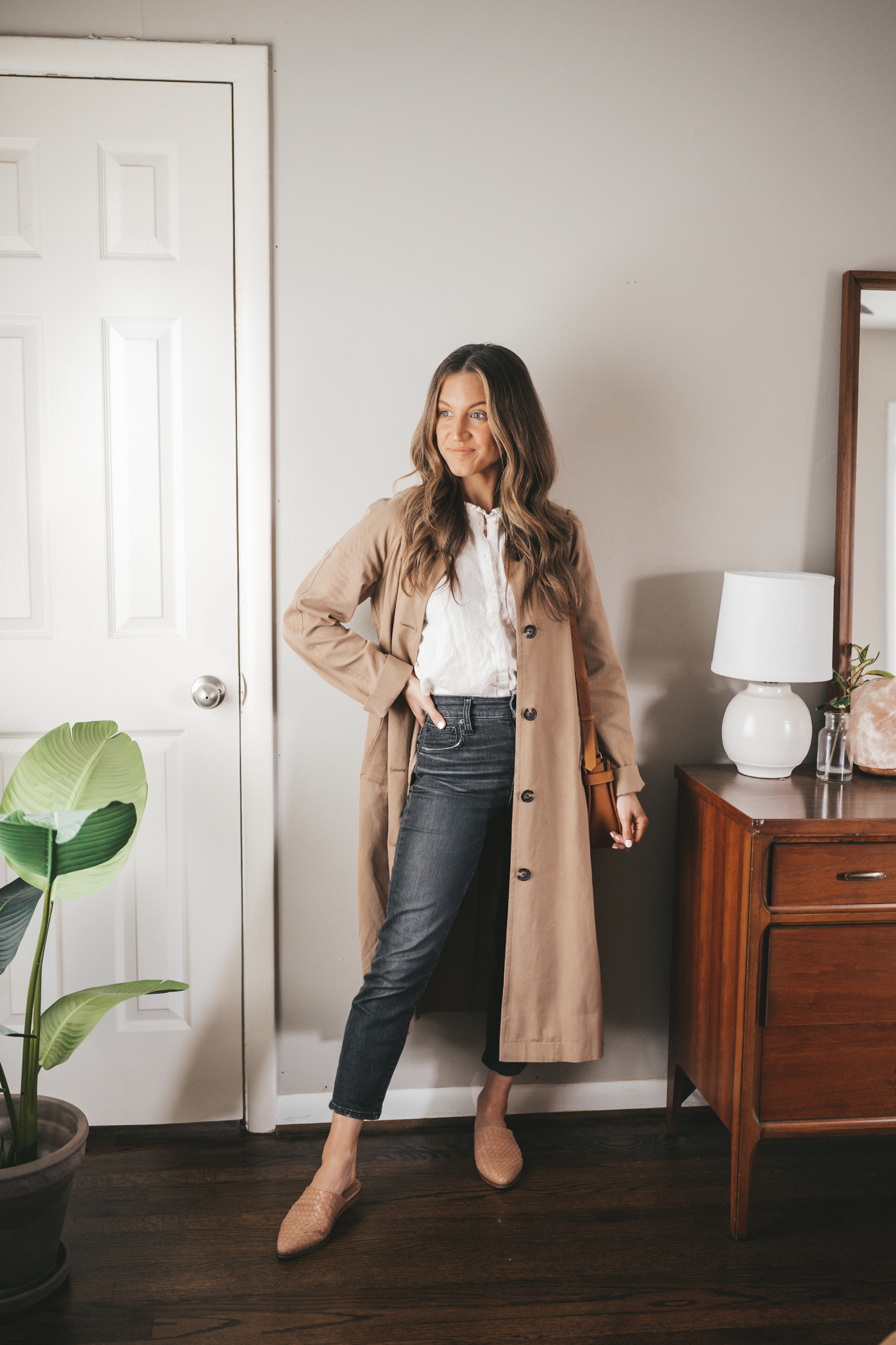 ashley wearing a white blouse, tan trench coat, washed black denim, and woven mules with a cognac crossbody bag