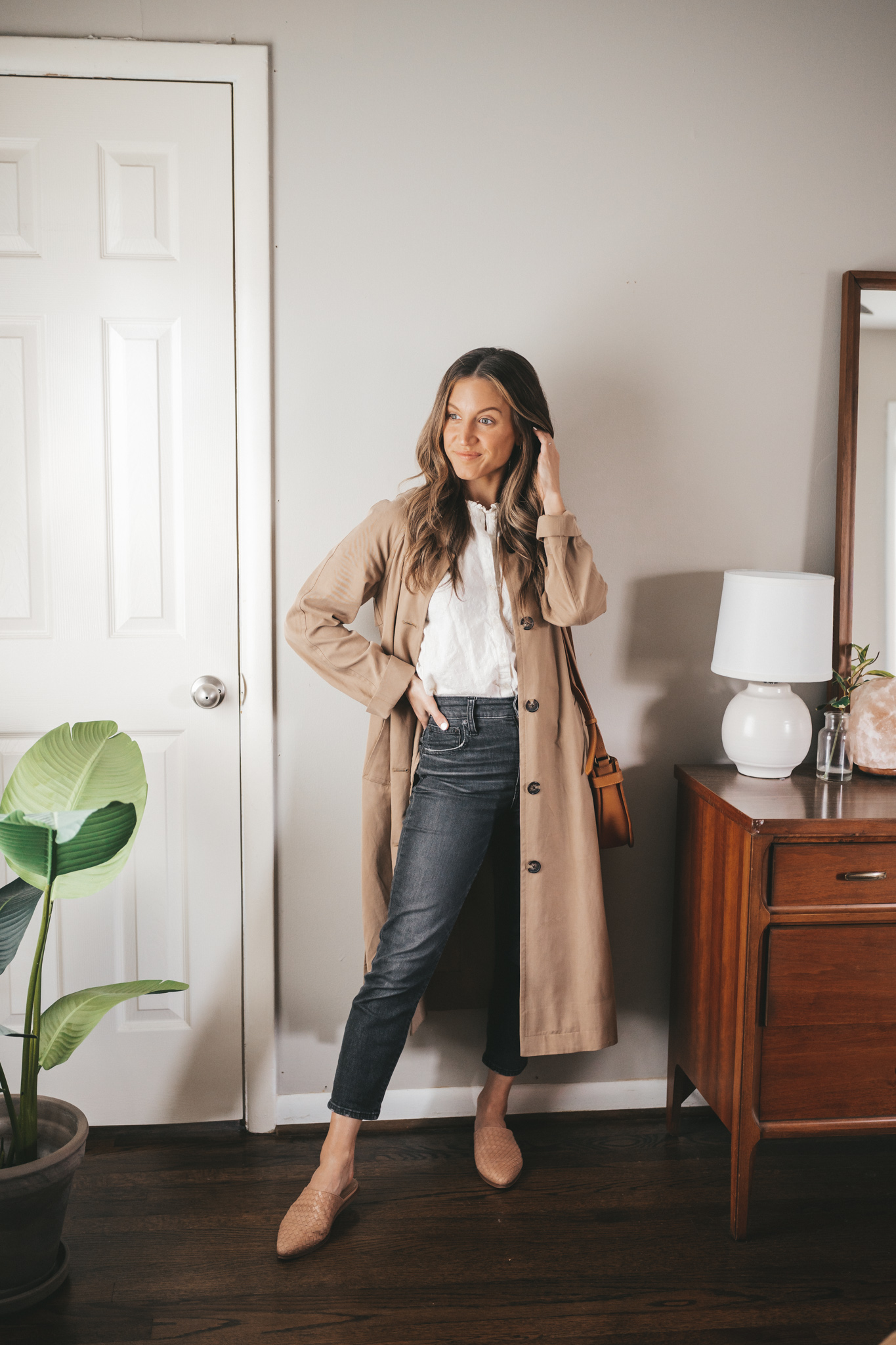 ashley wearing a white blouse, tan trench coat, washed black denim, and woven mules with a cognac crossbody bag