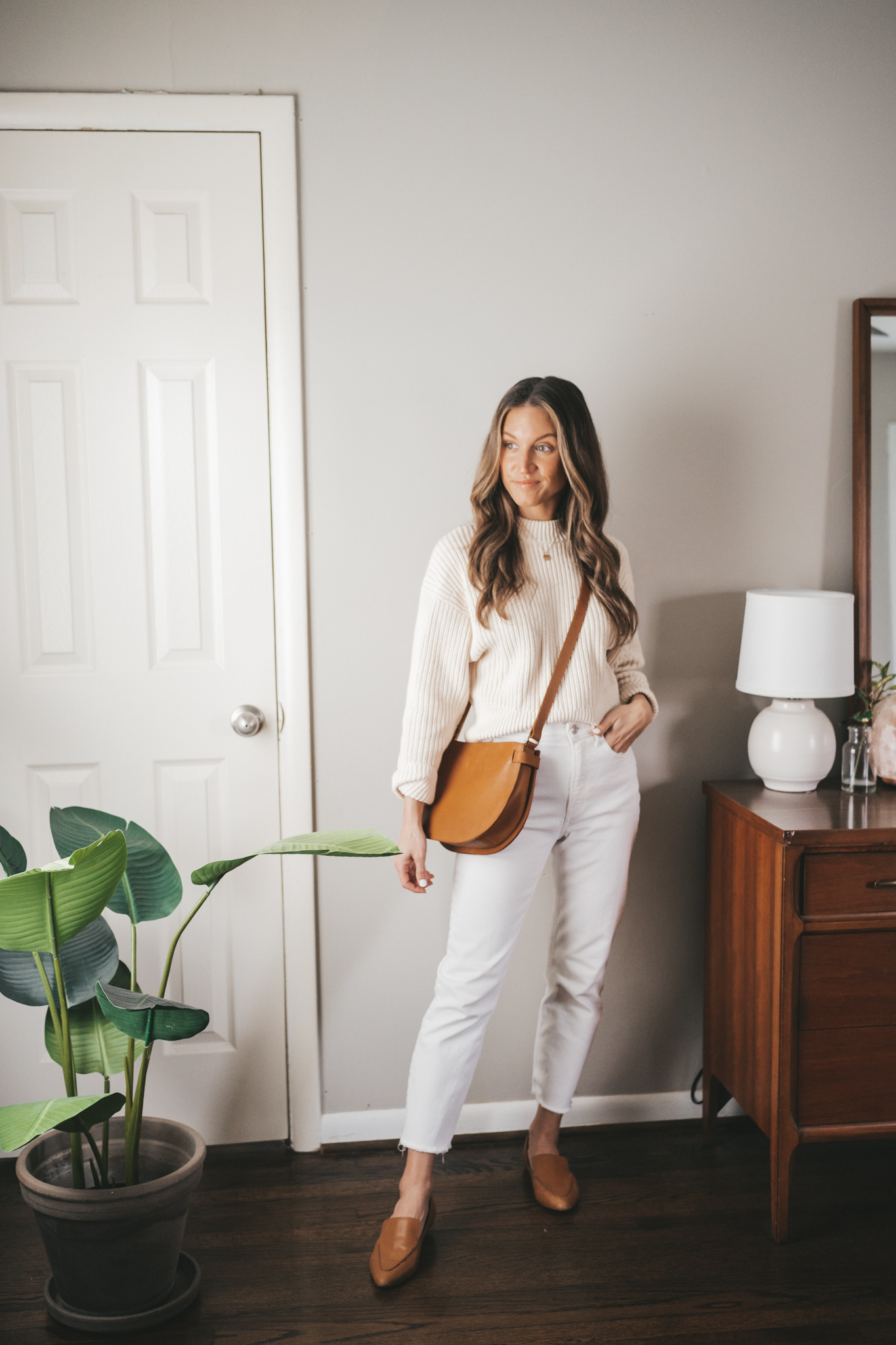 ashley wearing a cream colored sweater, white denim, cognac loafers, and a cognac crossbody bag