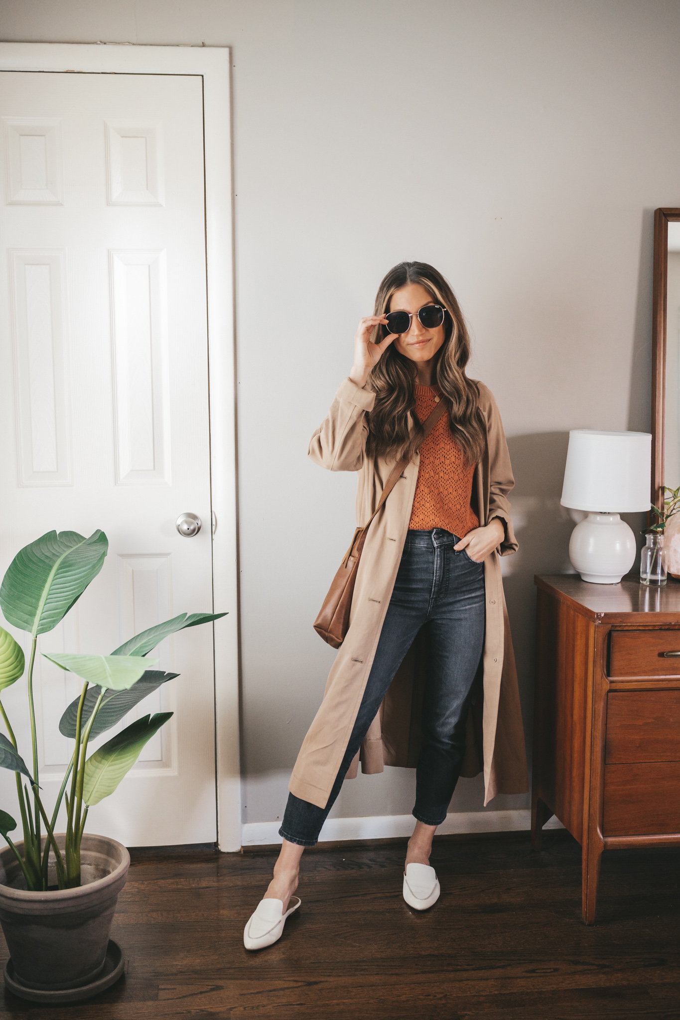 ashley wearing a rust colored sweater, tan trench coat, washed black denim, white mules, a brown crossbody bag, and oversized gold frame sunglasses